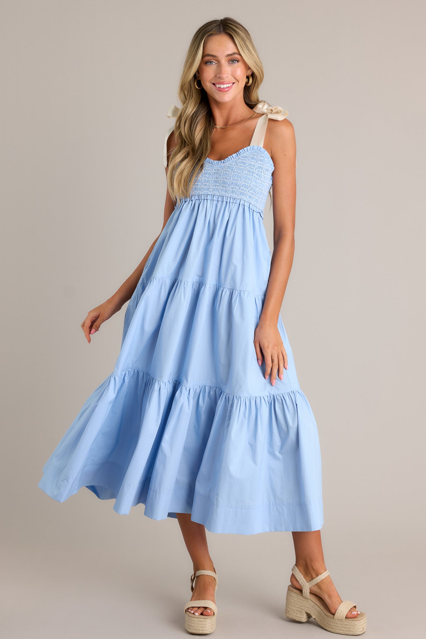 Front angled view of a light blue maxi dress featuring a v-neckline, thick self-tie straps, a fully smocked bodice, functional hip pockets, and a tiered design, w