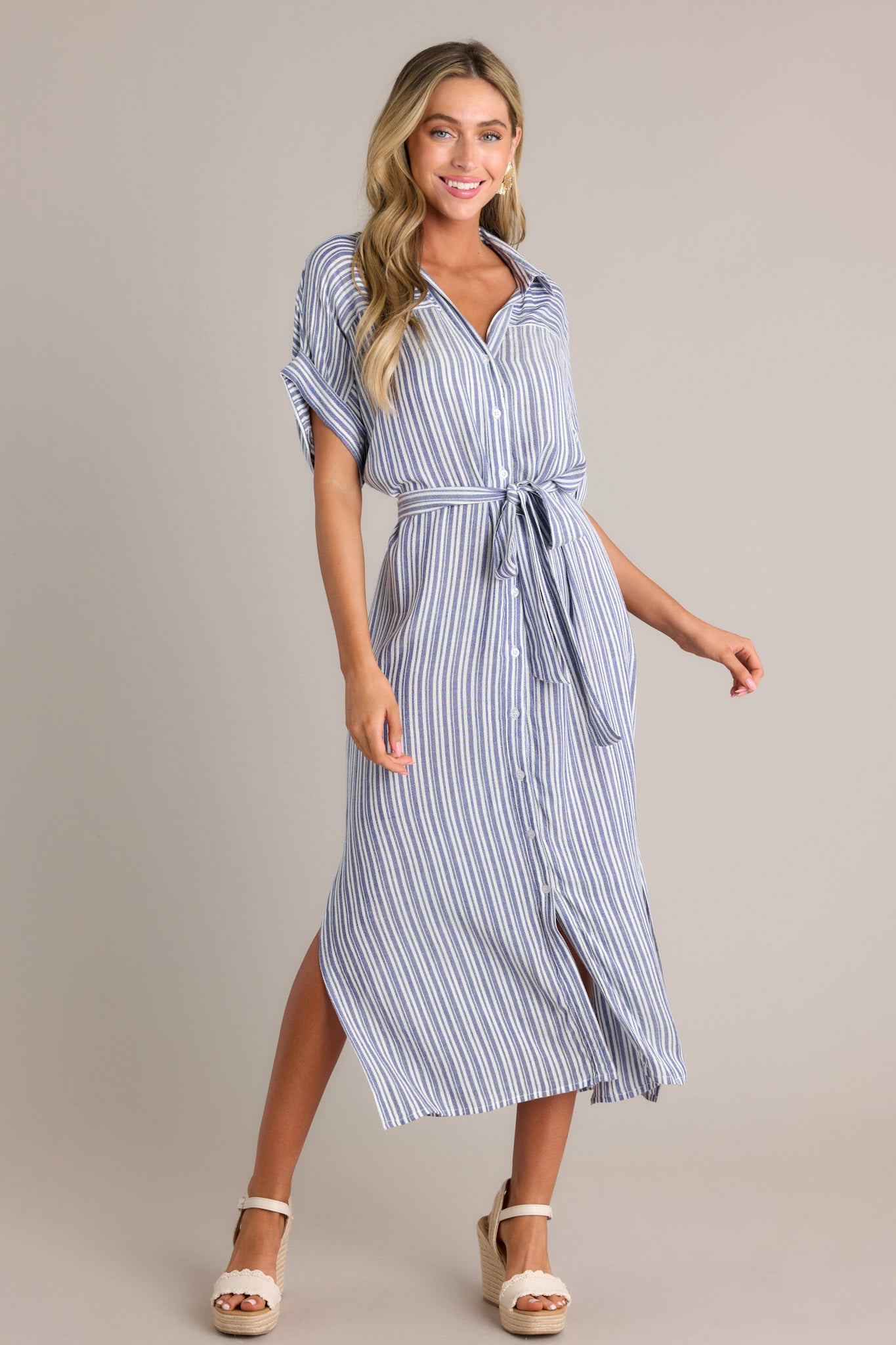 This blue stripe midi dress features a collared v-neckline, a functional button front, a self tie drawstring belt, functional pockets, side slits, and a flowing silhouette.