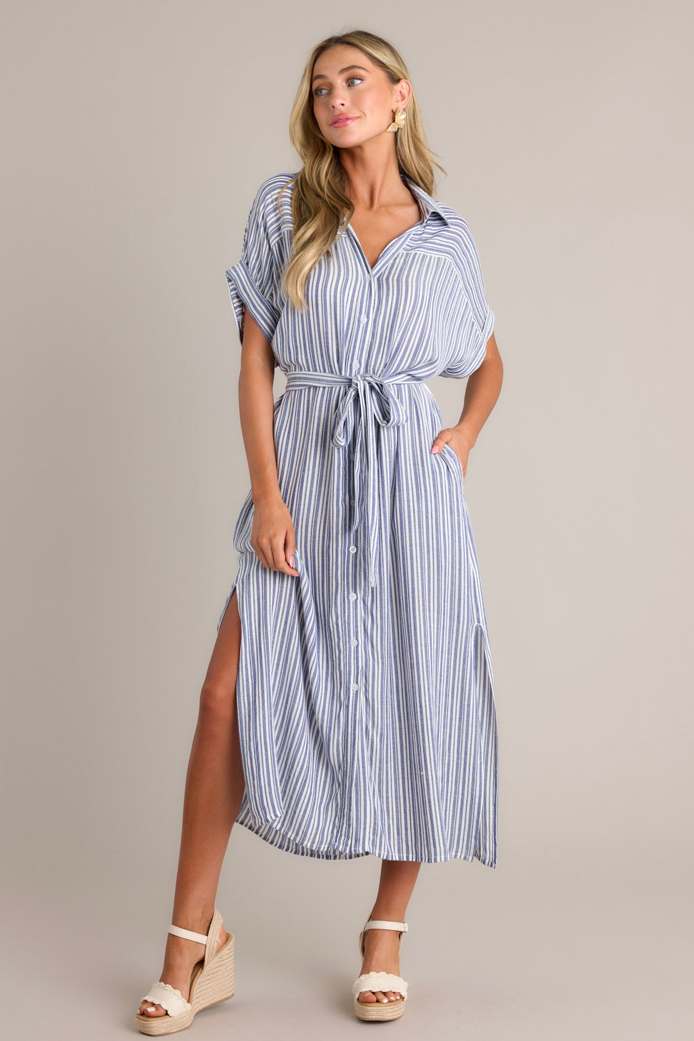 Front angled view of a blue stripe midi dress featuring a collared v-neckline, a functional button front, a self-tie drawstring belt, functional pockets, side slits, and a flowing silhouette