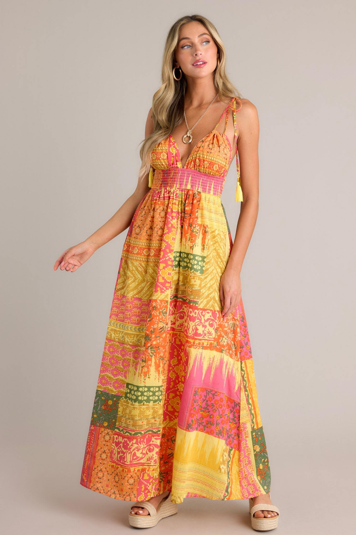 Full length view of an orange maxi dress with a v-neckline, adjustable self-tie straps, a low back, a fully smocked waistband, a unique patchwork design, and a flowing silhouette