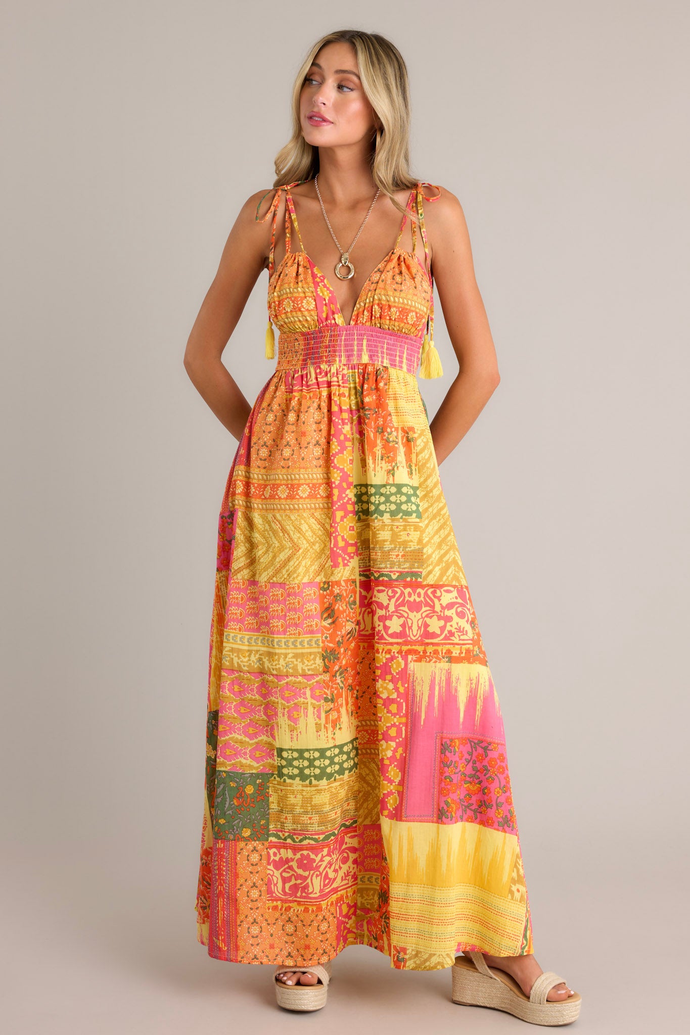 Front view of an orange maxi dress featuring a v-neckline, adjustable self-tie straps, a fully smocked waistband, a unique patchwork design, and a flowing silhouette.