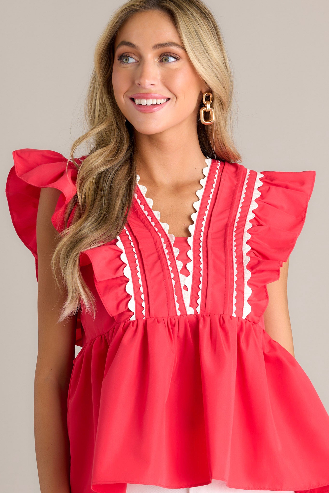 Front angled view of a red babydoll top featuring a v-neckline, scalloped & ruffled bodice detailing, a discrete side zipper, and short flutter sleeves