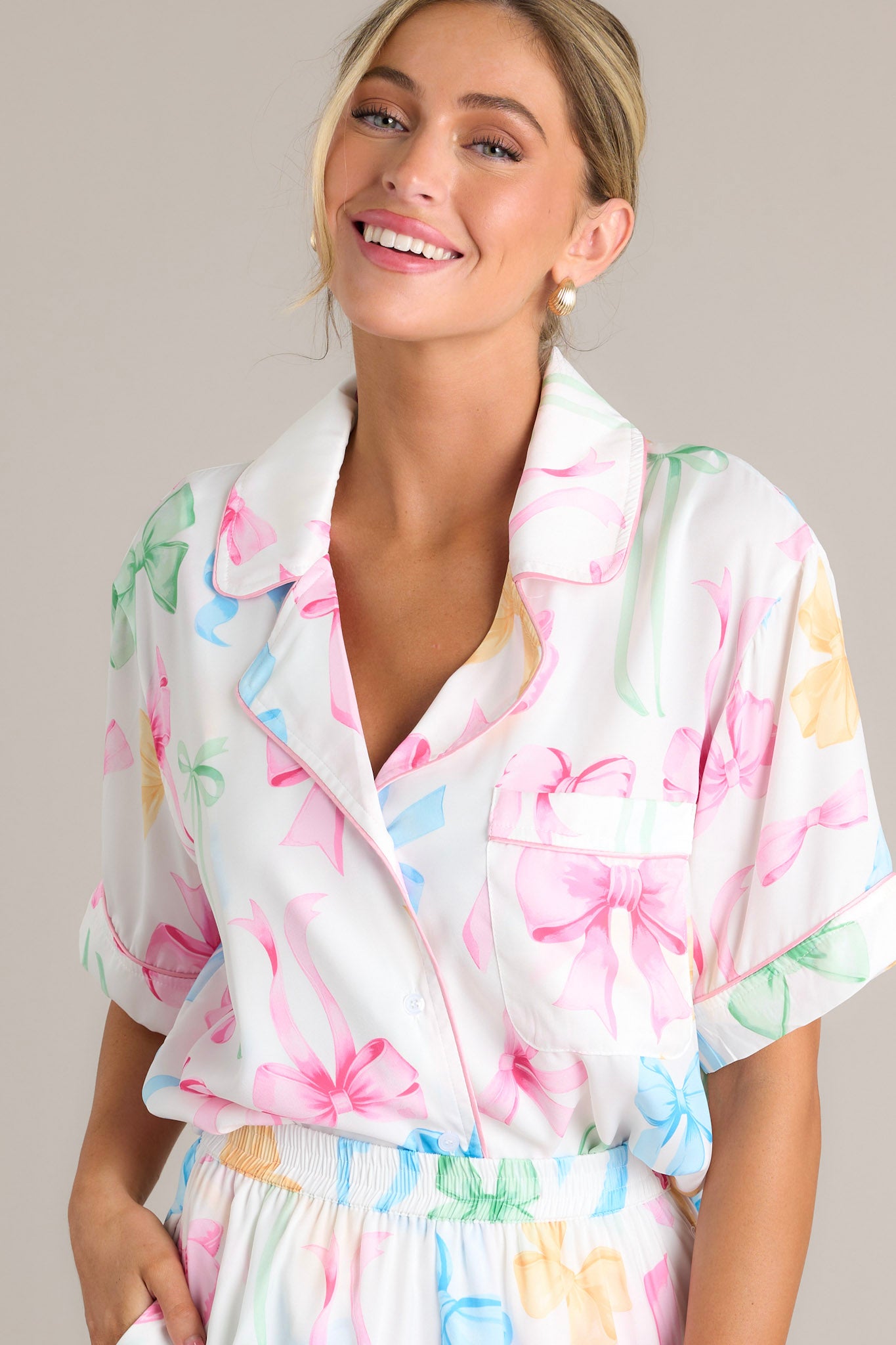 Front angled view of a white pajama top featuring a notched lapel collared neckline, a chest pocket, a full button front, a fun multi-color bow print, and a split hemline