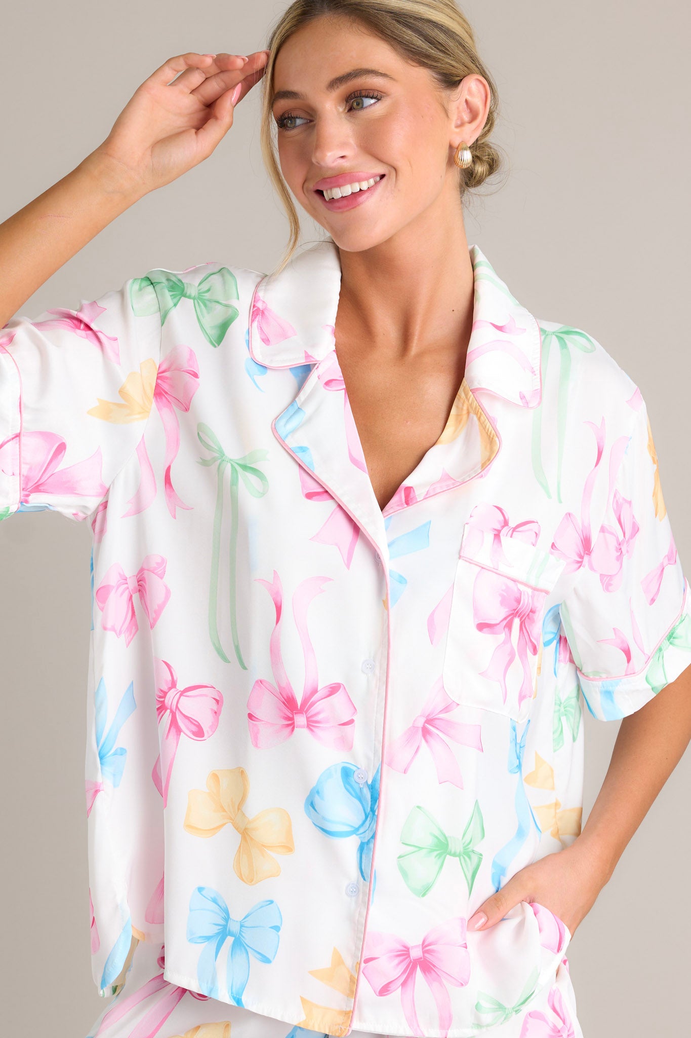 Front view of a white pajama top featuring a notched lapel collared neckline, a chest pocket, a full button front, a fun multi-color bow print, and a split hemline.