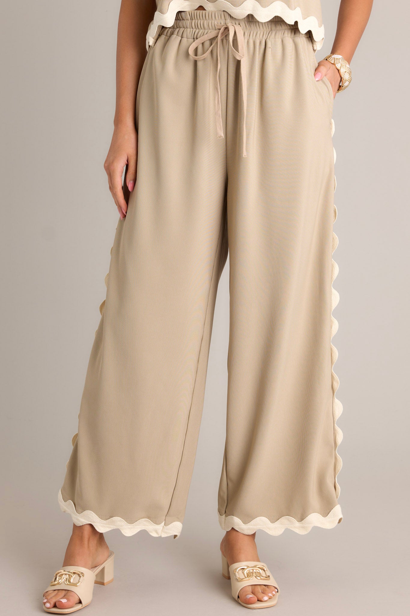 Front view of taupe pants featuring a high waisted design, an elastic waistband, a self-tie drawstring, functional hip pockets, a ribbed texture, and ricrac hemlines.