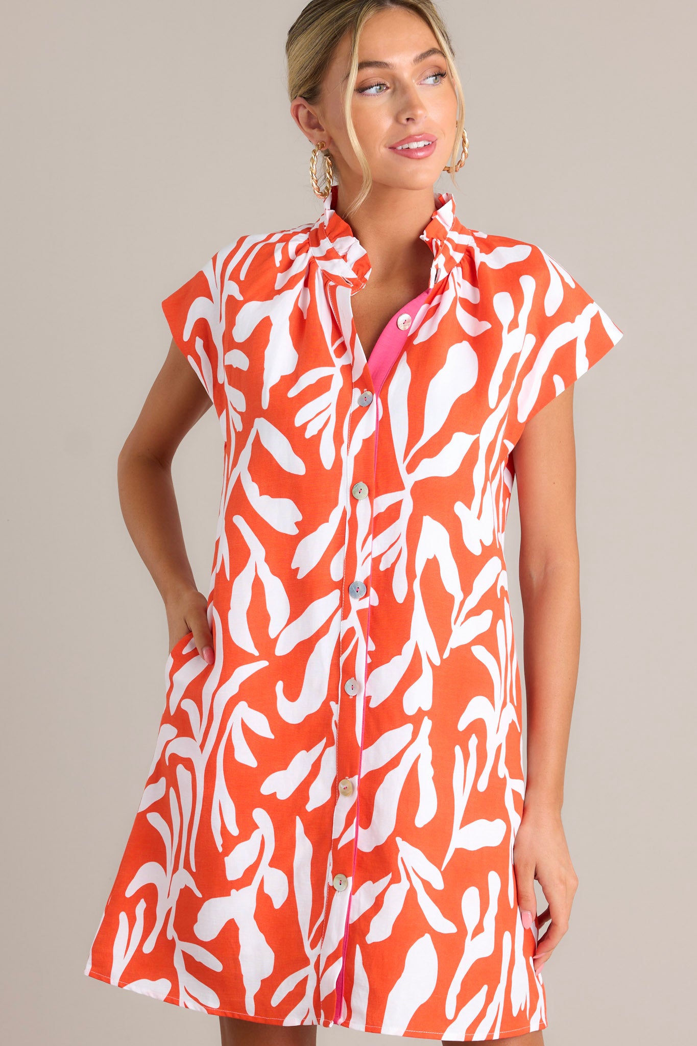 Front view of a red orange dress featuring a ruffled collar, caped short sleeves, functional buttons, a fun abstract print, and functional hip pockets.