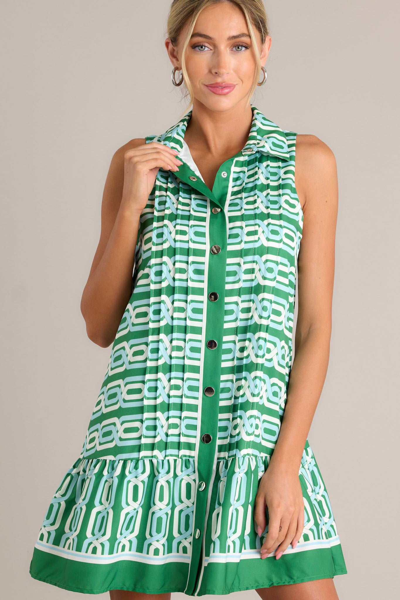 Front view of a green multi mini dress featuring a collared neckline, a functional button front, pleats, a unique pattern, a single tier, and a sleeveless design.