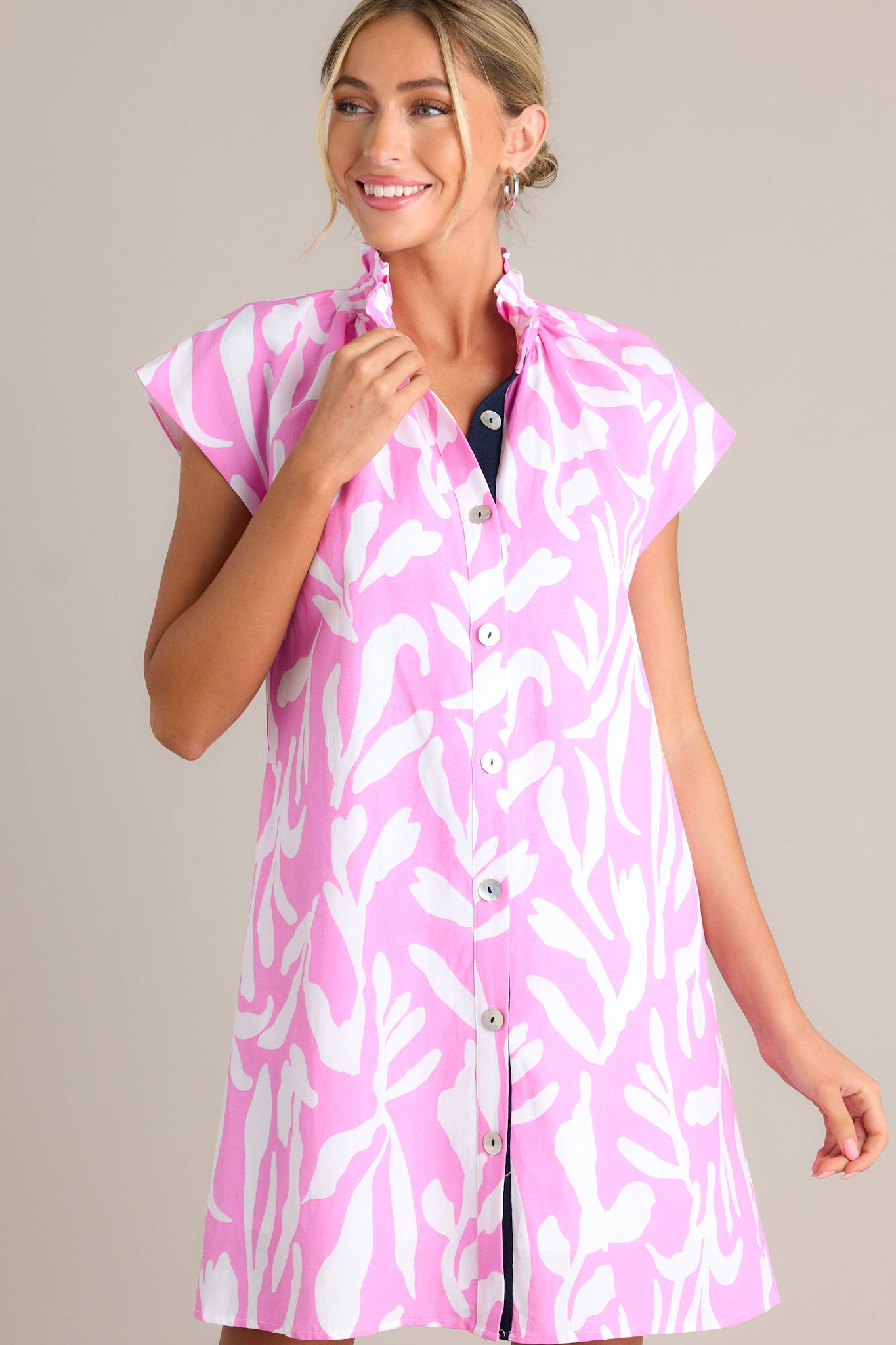 Front angled view of a pink dress featuring a ruffled collar, caped short sleeves, functional buttons, a fun abstract print, and functional hip pockets