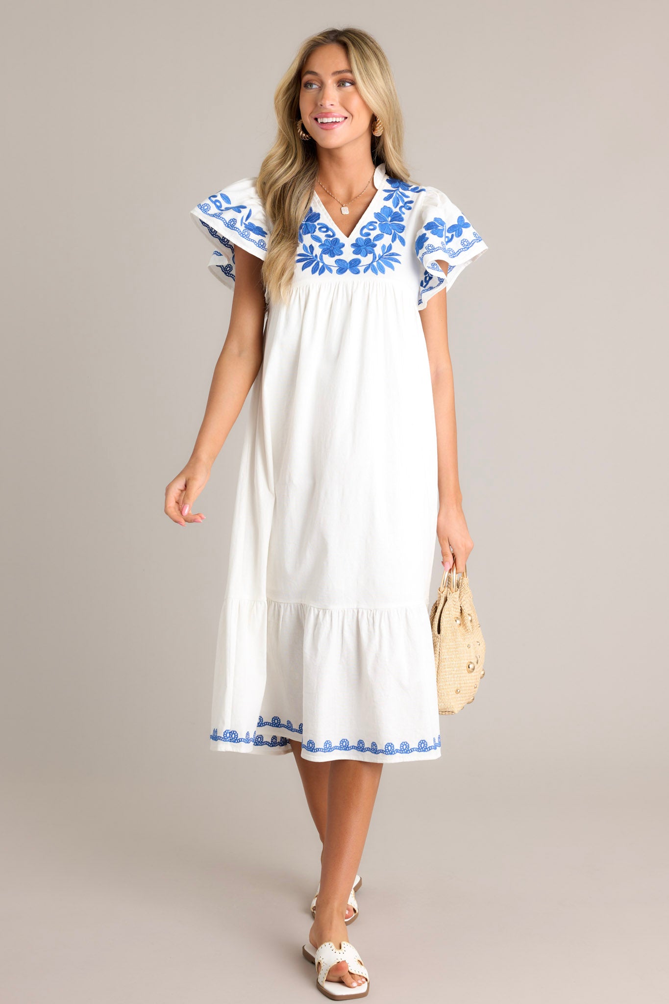 This ivory midi dress features a ruffled v-neckline, blue floral embroidery, a single tier, an embroidered hemline, and short embroidered flutter sleeves.