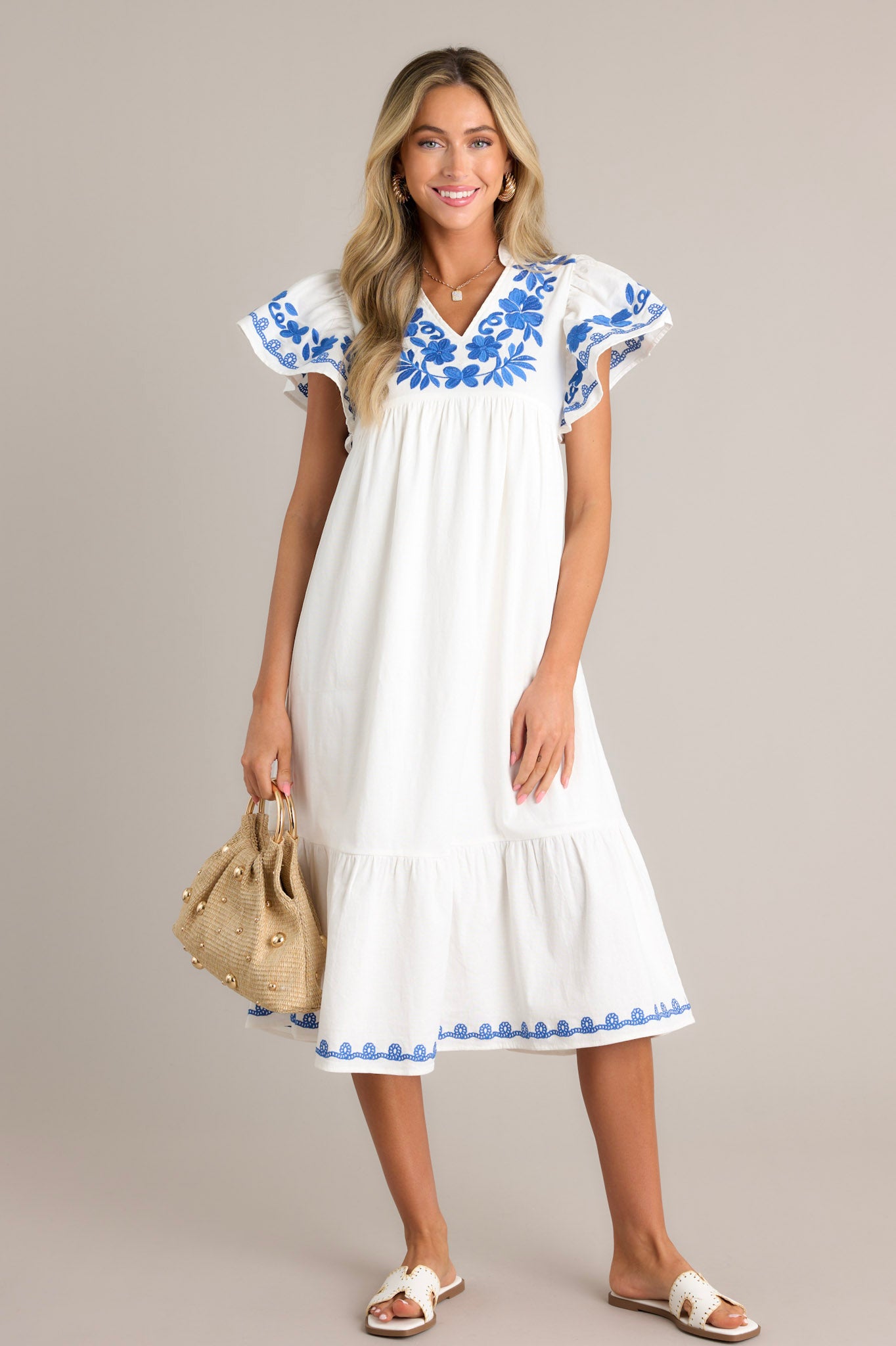 Full length view of an ivory midi dress with a ruffled v-neckline, blue floral embroidery, a single tier, an embroidered hemline, and short embroidered flutter sleeves