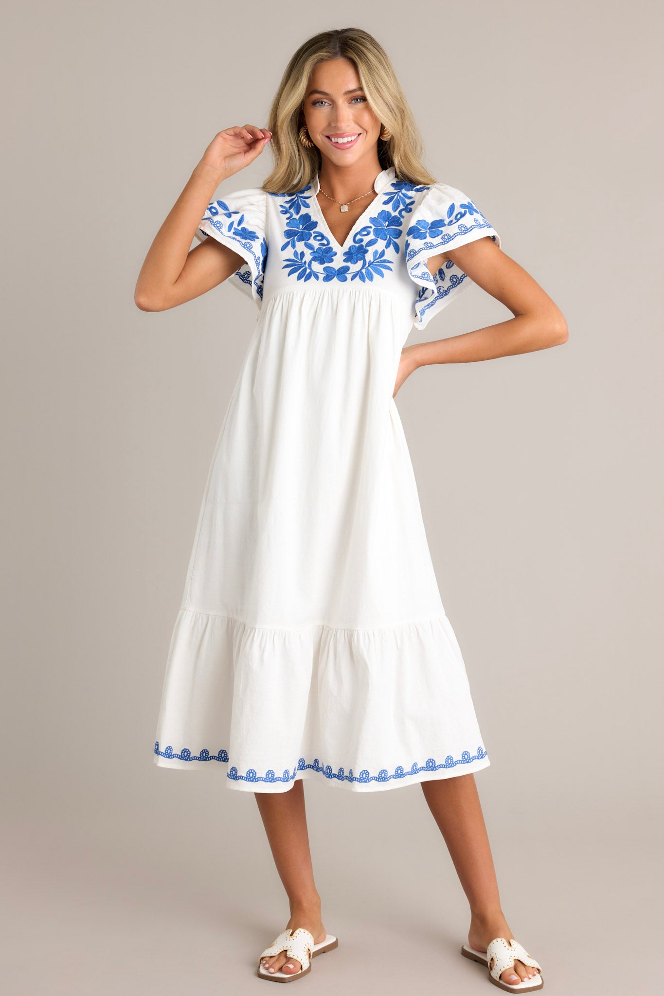 Front view of an ivory midi dress featuring a ruffled v-neckline, blue floral embroidery, a single tier, an embroidered hemline, and short embroidered flutter sleeves.