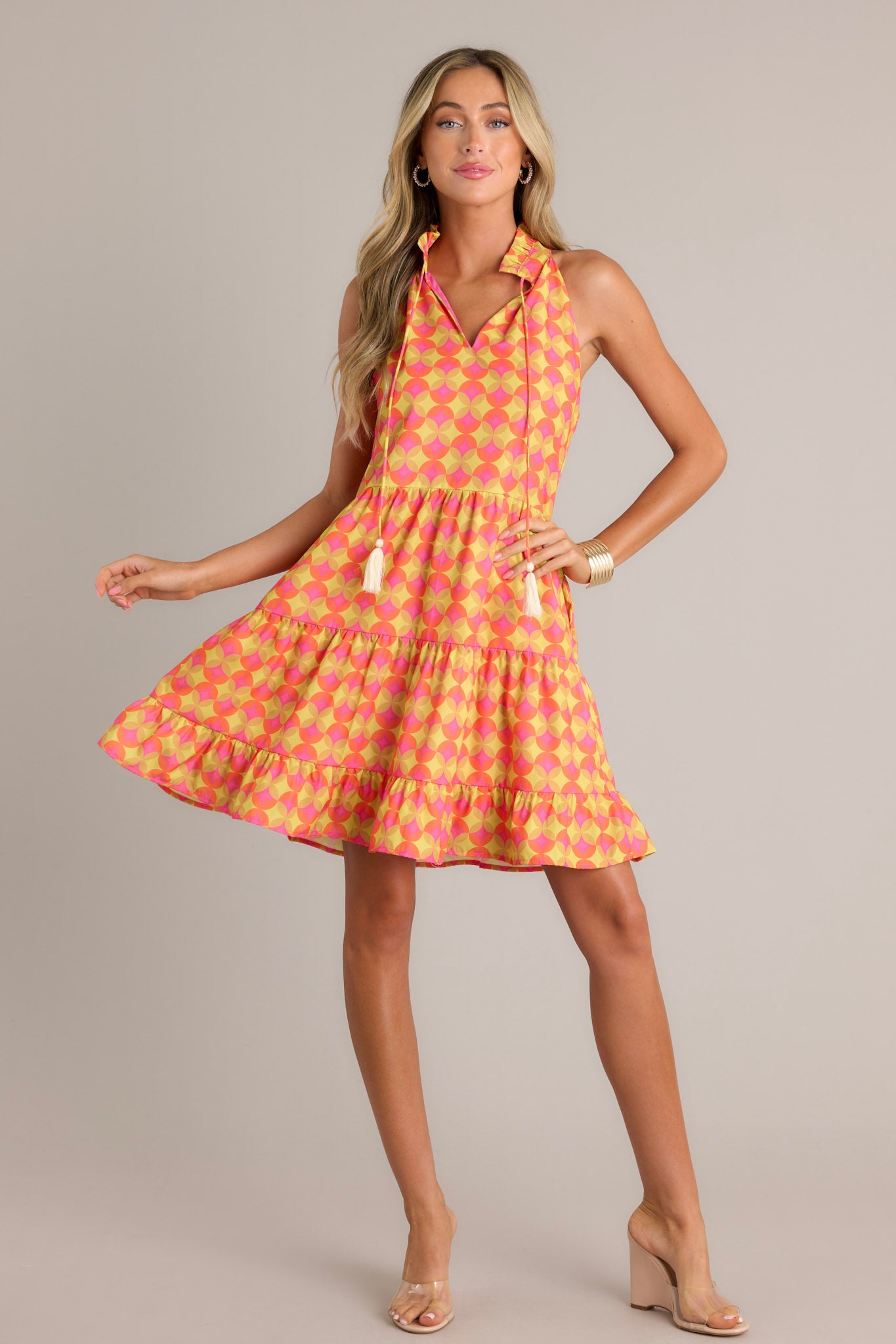 Full length view of an orange mini dress with a ruffled v-neckline, a self-tie neck feature, a unique pattern, functional hip pockets, multiple tiers, and a flowing silhouette