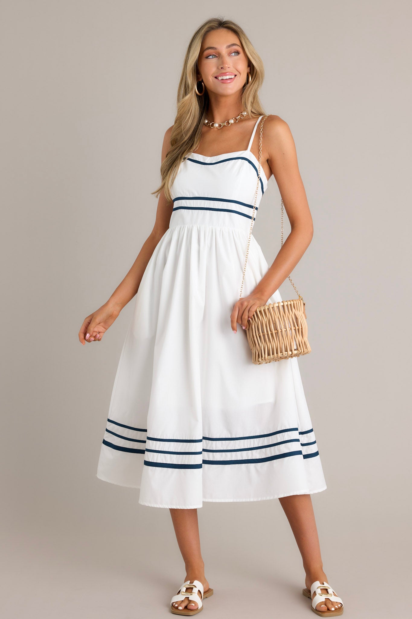 Full length view of an ivory midi dress with a slight sweetheart neckline, navy trim, thin adjustable straps, a smocked back insert, a navy striped waist, functional hip pockets, and a navy striped hemline