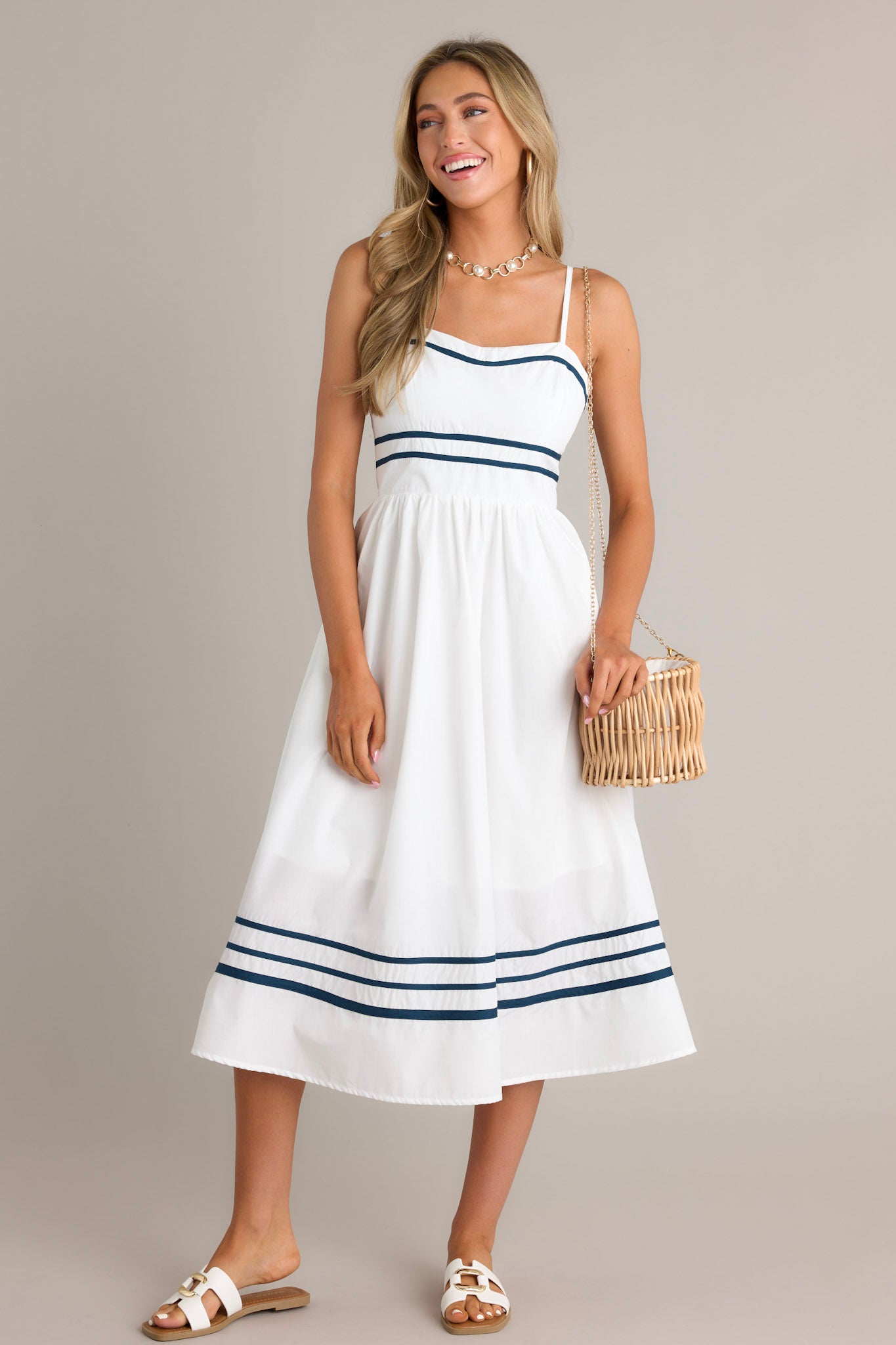 Angled Full length view of an ivory midi dress with a slight sweetheart neckline, navy trim, thin adjustable straps, a smocked back insert, a navy striped waist, functional hip pockets, and a navy striped hemline