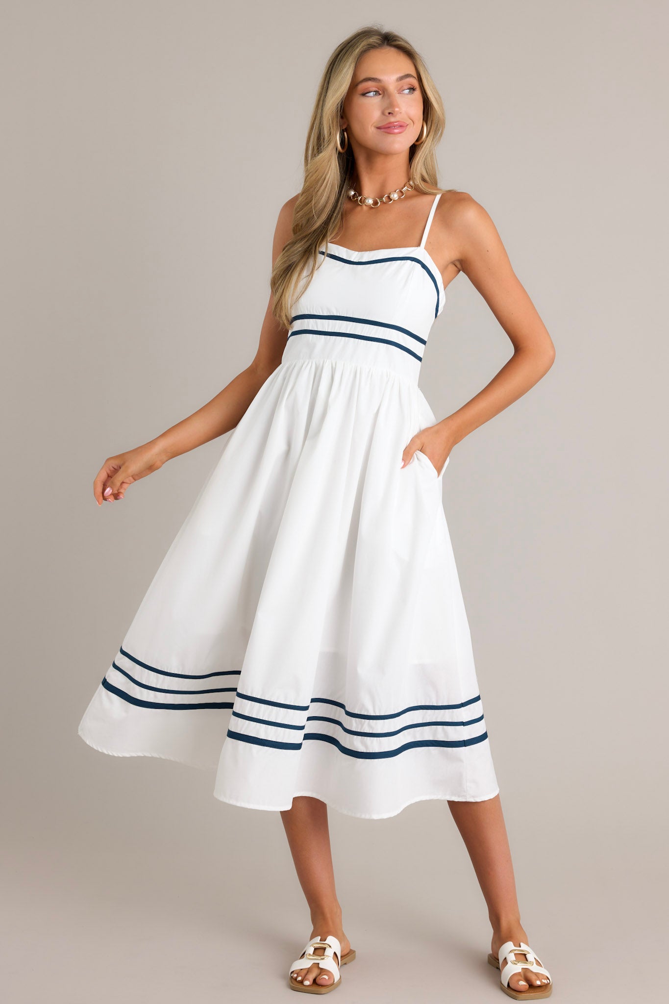 Action shot of an ivory midi dress displaying the fit and movement, highlighting the slight sweetheart neckline, navy trim, thin adjustable straps, functional hip pockets, and navy striped hemline.