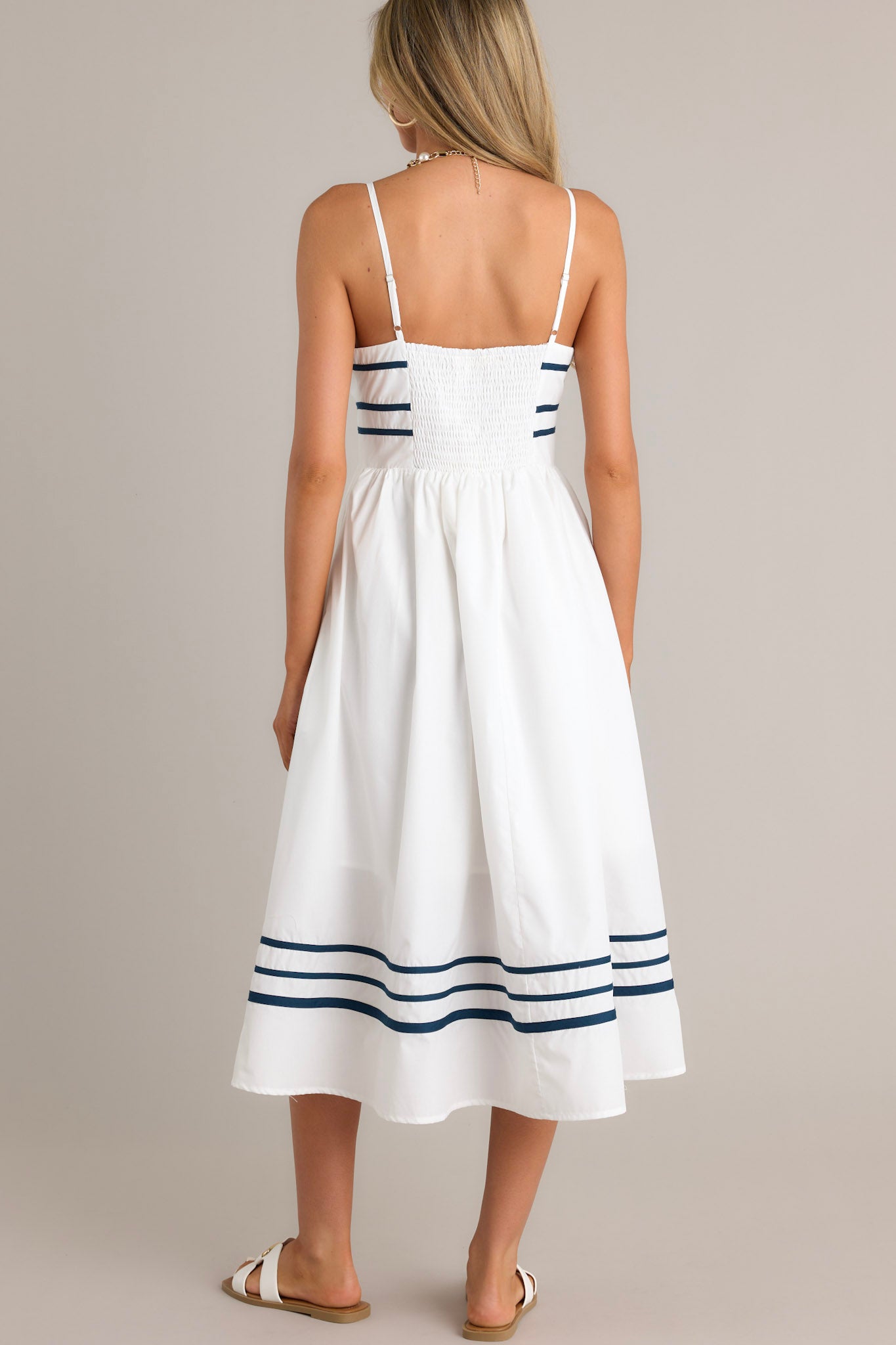 Back view of an ivory midi dress highlighting the smocked back insert, navy trim, and the navy striped hemline.