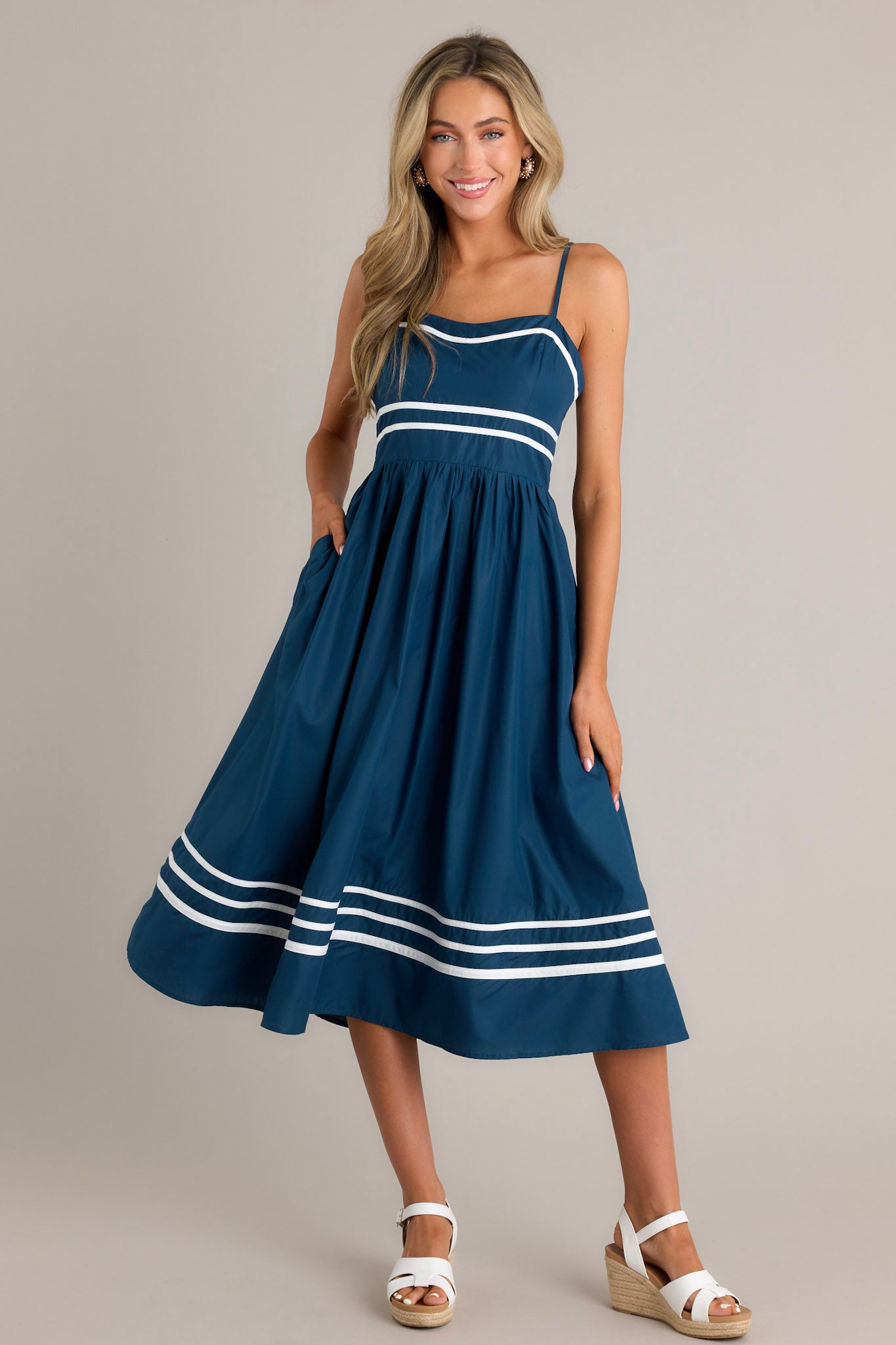 This navy midi dress features a slight sweetheart neckline, ivory trim, thin adjustable straps, a smocked back insert, an ivory striped waist, functional hip pockets, and an ivory striped hemline.