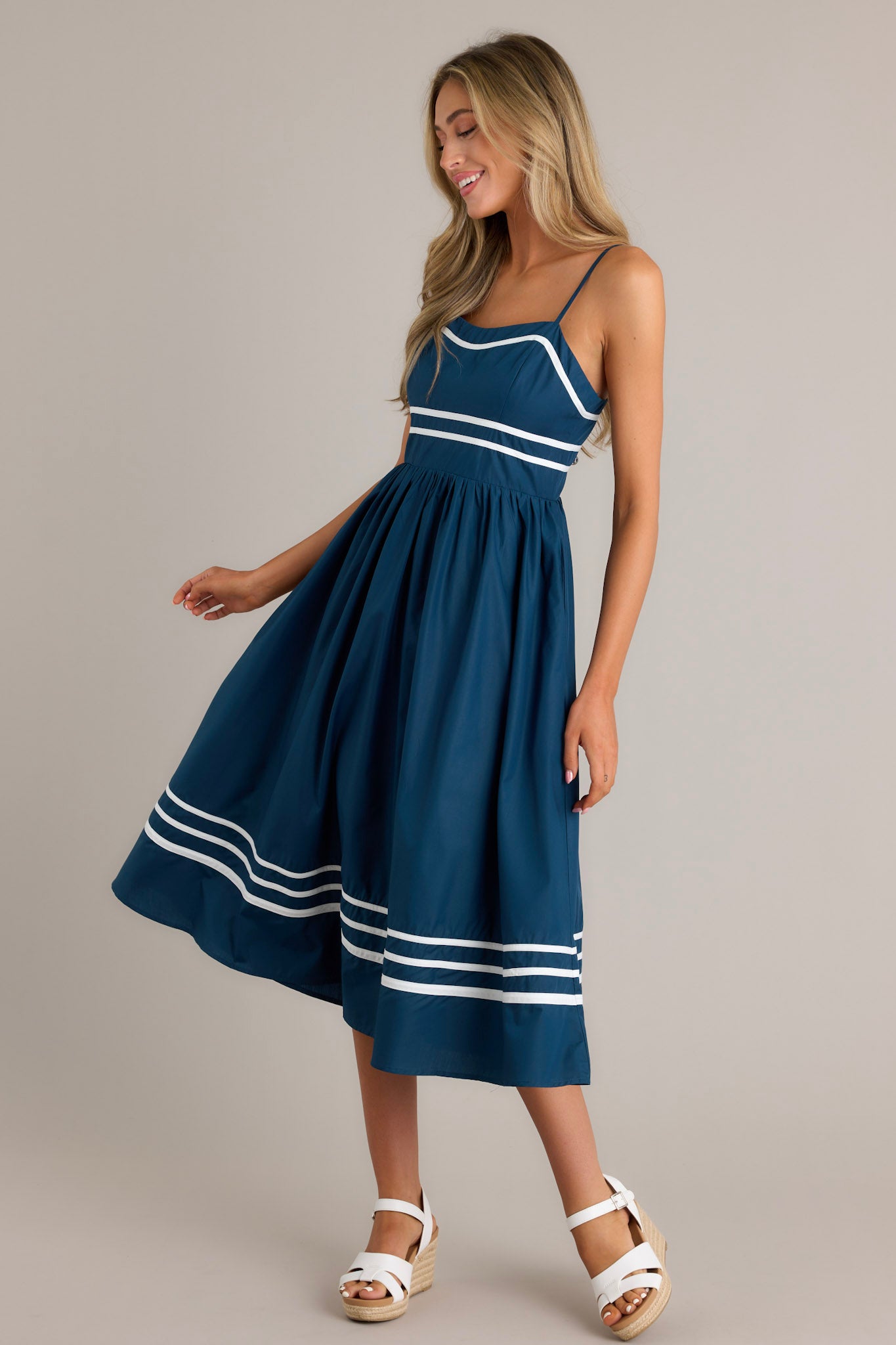 Side view of a navy midi dress showcasing the slight sweetheart neckline, ivory trim, thin adjustable straps, functional hip pockets, and an ivory striped hemline.  Back View: