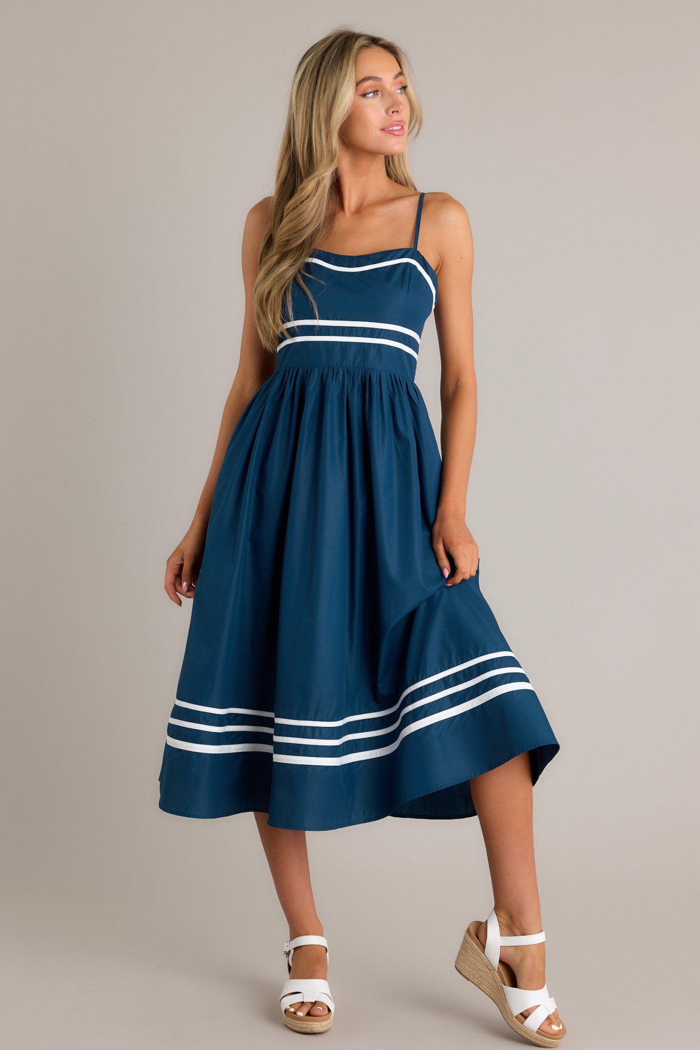 Front angled view of a navy midi dress featuring a slight sweetheart neckline, ivory trim, thin adjustable straps, a smocked back insert, an ivory striped waist, functional hip pockets, and an ivory striped hemline