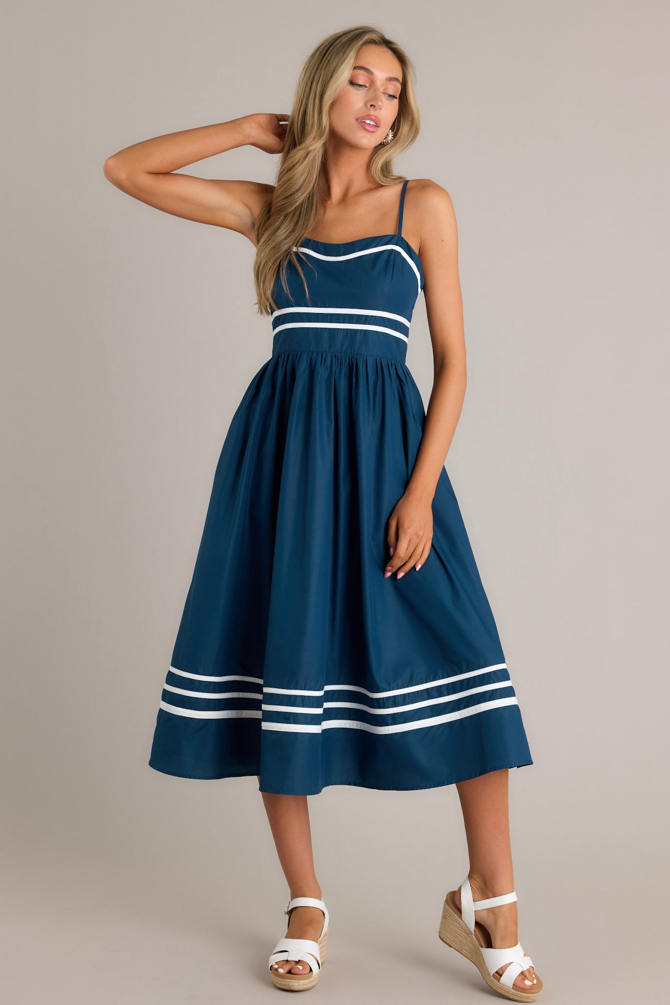 Full length view of a navy midi dress with a slight sweetheart neckline, ivory trim, thin adjustable straps, a smocked back insert, an ivory striped waist, functional hip pockets, and an ivory striped hemline