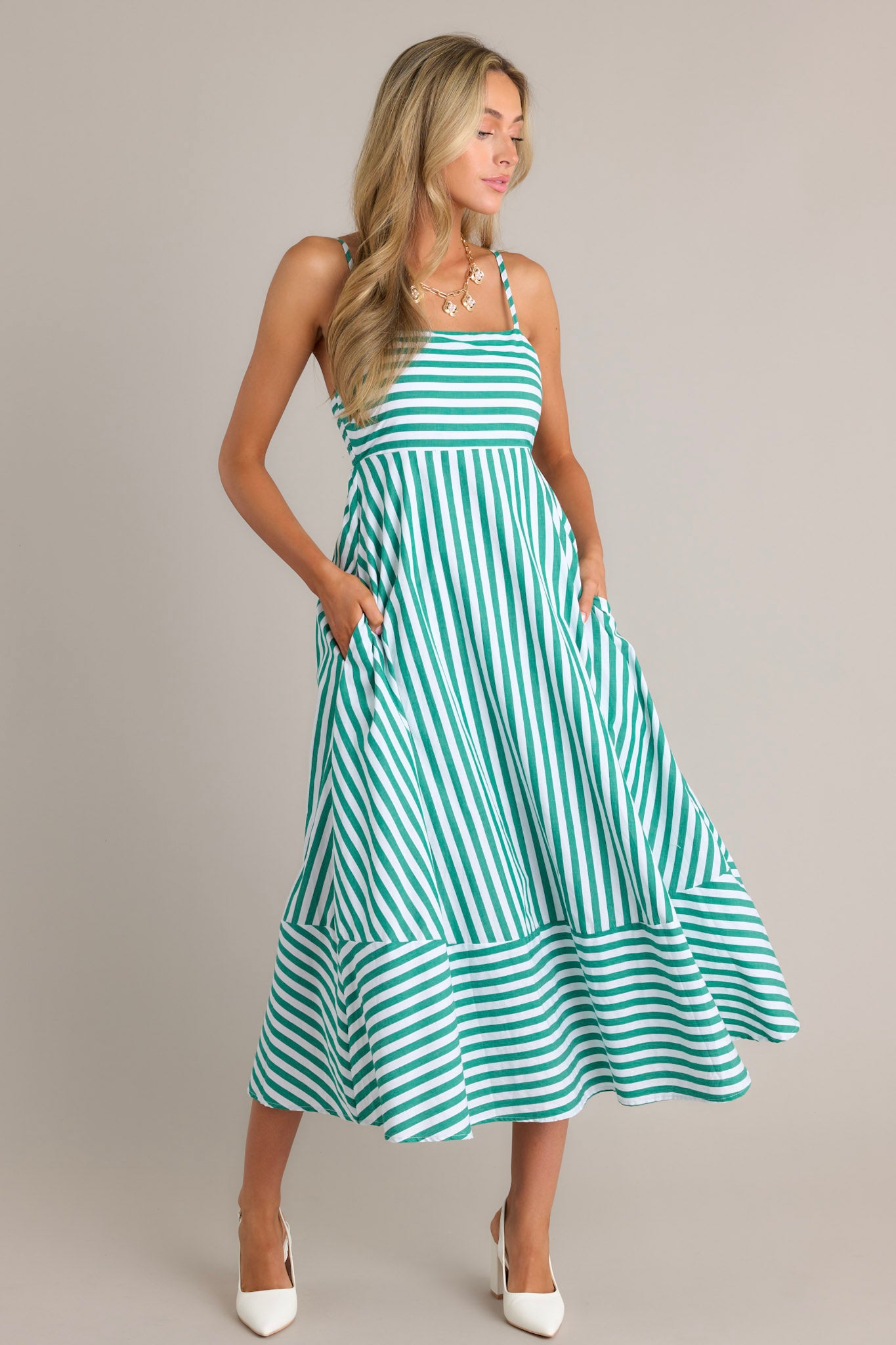 Front angled view of a green stripe midi dress featuring a square neckline, thin adjustable straps, a smocked back insert, functional hip pockets, and horizontal & vertical stripes