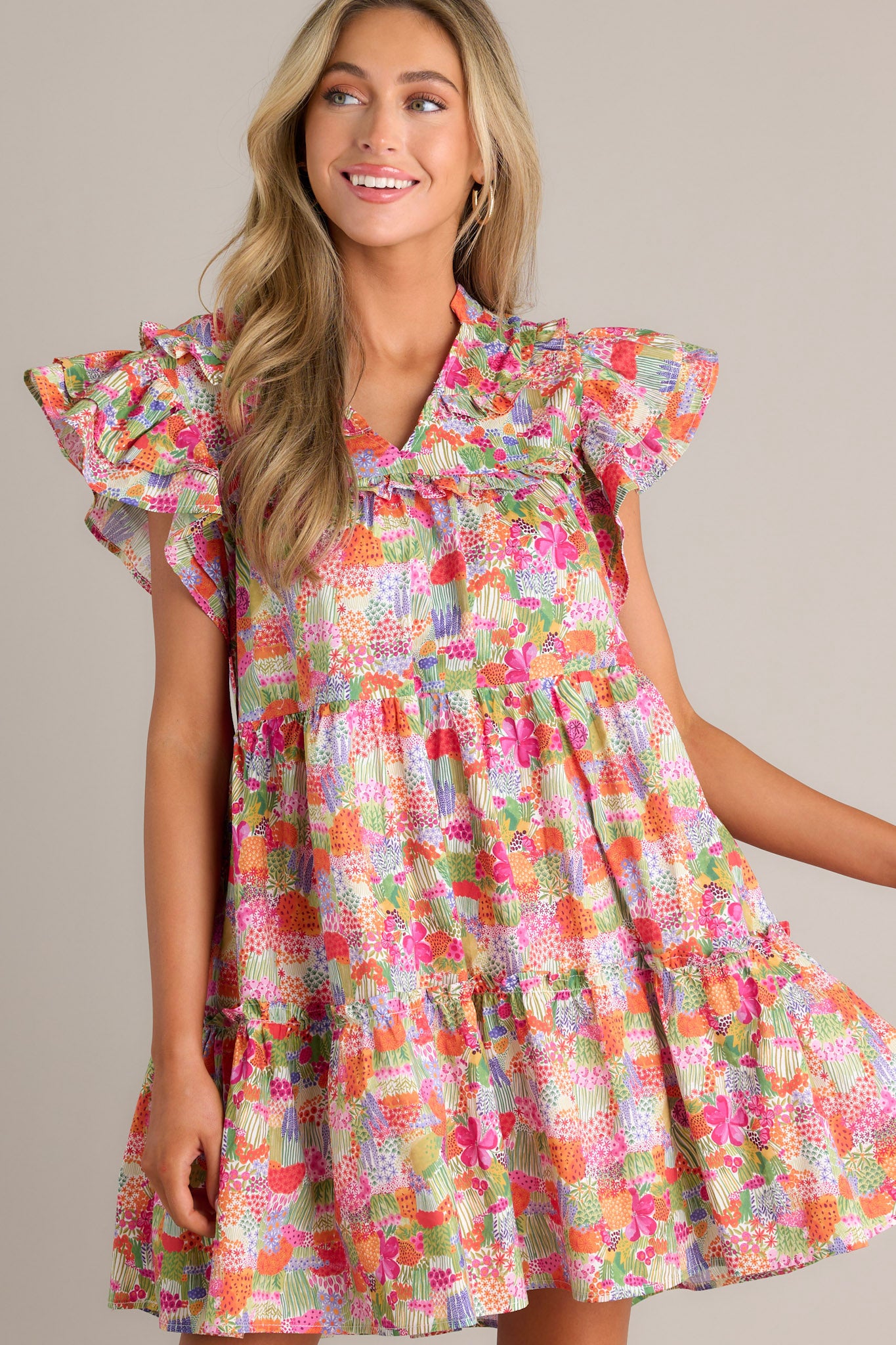 Front angled view of a pink mini dress featuring a v-neckline, ruffles on the chest, a tiered design, functional hip pockets, and short flutter sleeves