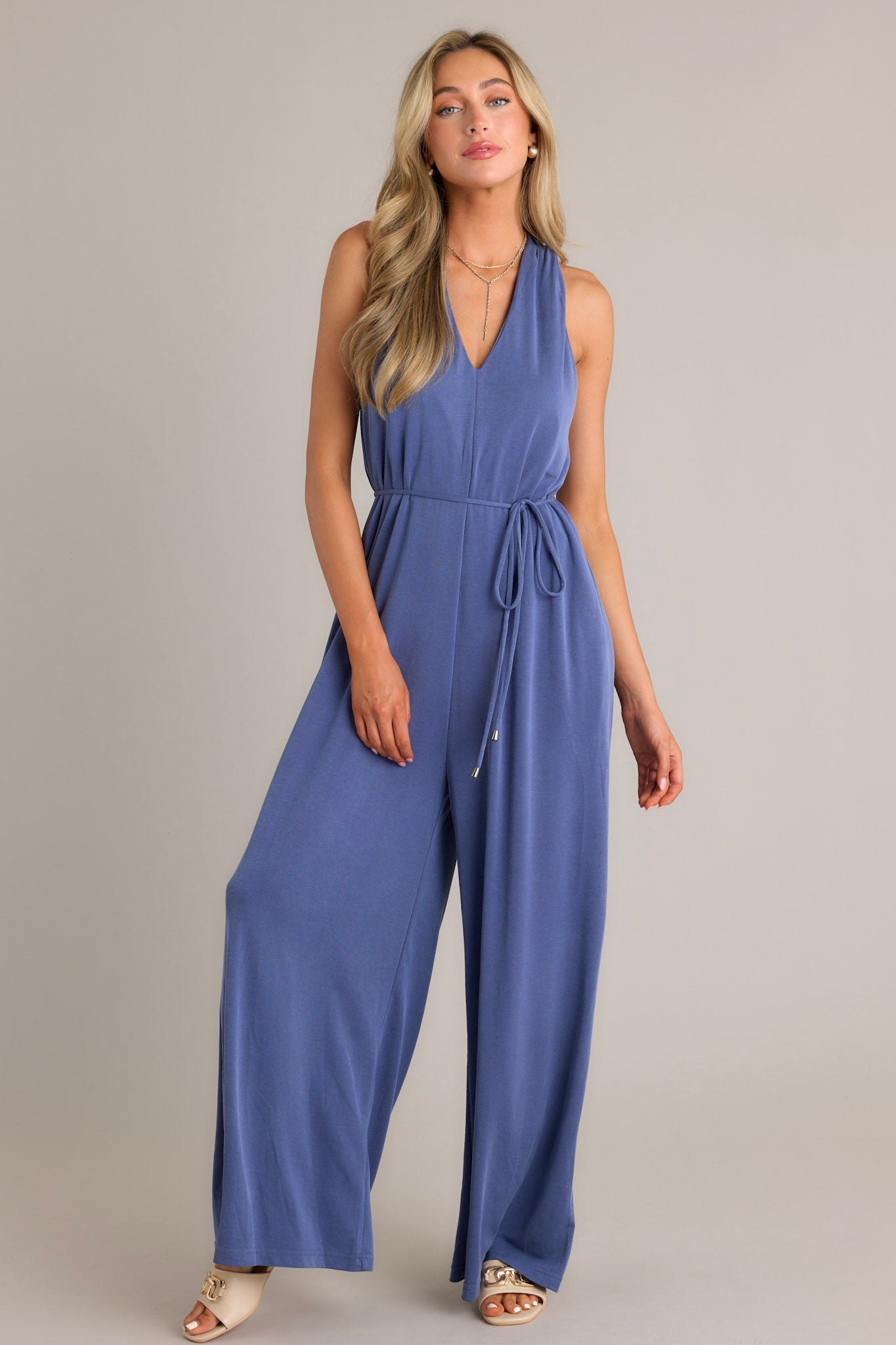 Front view of a violet blue jumpsuit featuring a v-neckline, a self-tie waist belt, functional hip pockets, a wide leg design, and a racerback.