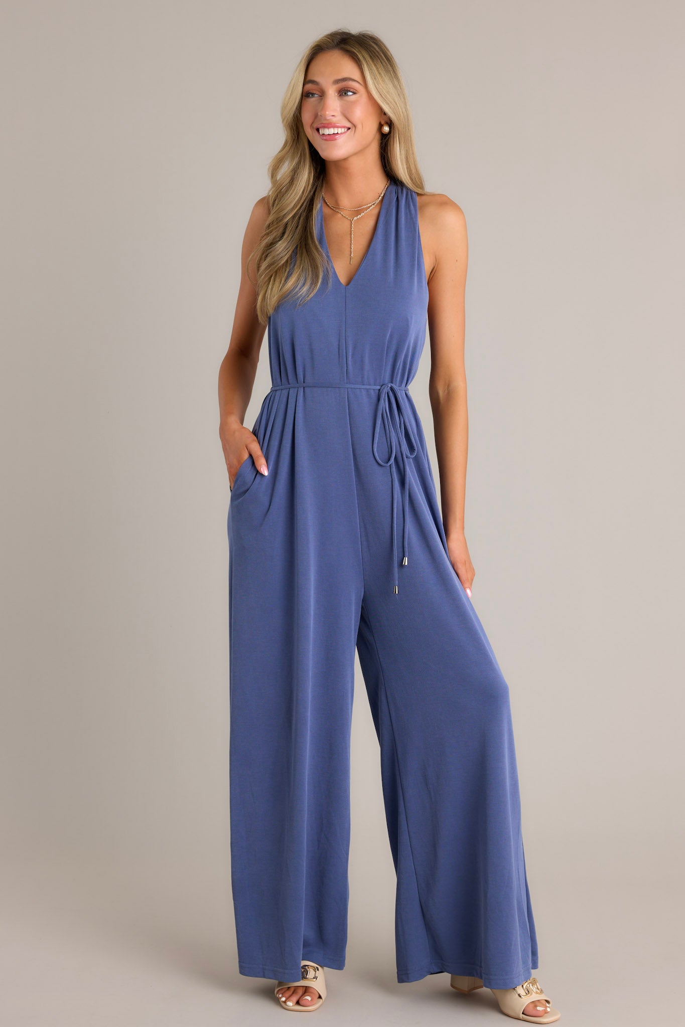 Front angled view of a violet blue jumpsuit featuring a v-neckline, a self-tie waist belt, functional hip pockets, a wide leg design, and a racerback