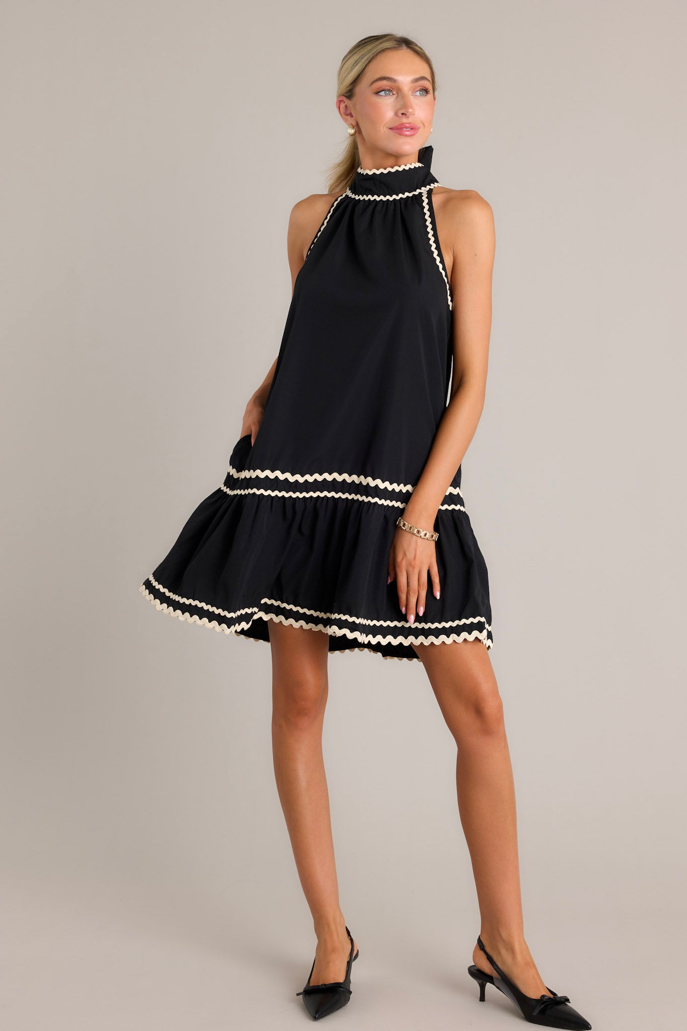 This black midi dress features a high self-tie neckline with a keyhole cutout, ivory ricrac detailing throughout, functional hip pockets, a single tier, a ricrac hemline, and a sleeveless design.