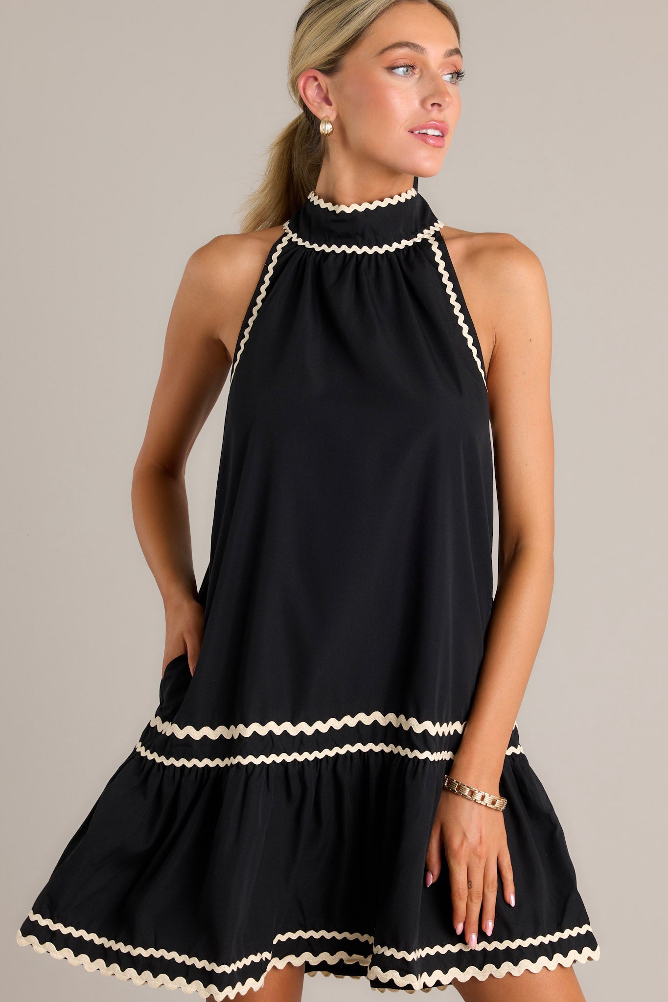 Front angled view of a black midi dress featuring a high self-tie neckline with a keyhole cutout, ivory ricrac detailing throughout, functional hip pockets, a single tier, a ricrac hemline, and a sleeveless design