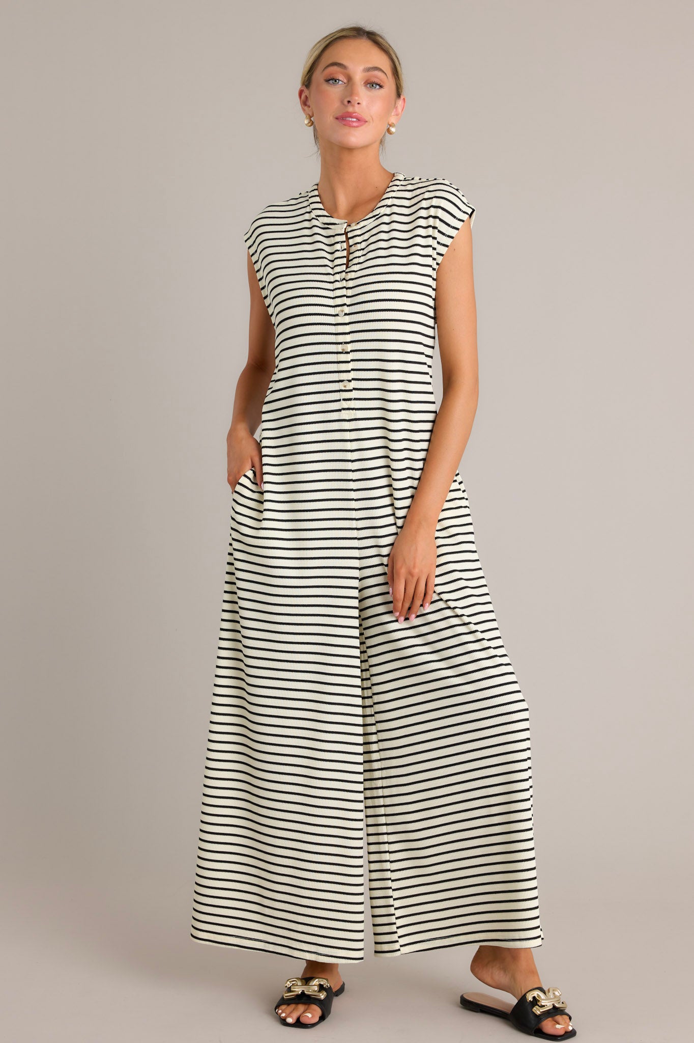 Full body view of a black striped jumpsuit with a rounded neckline, functional button front bodice, functional hip pockets, a textured material, and a sleeveless design