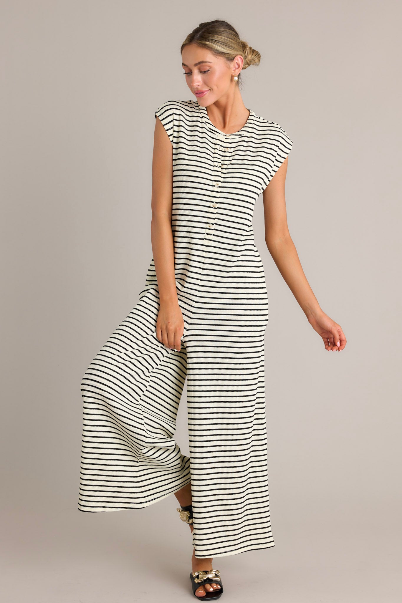 Action shot of a black striped jumpsuit displaying the fit and movement, highlighting the rounded neckline, functional button front bodice, functional hip pockets, textured material, and sleeveless design.