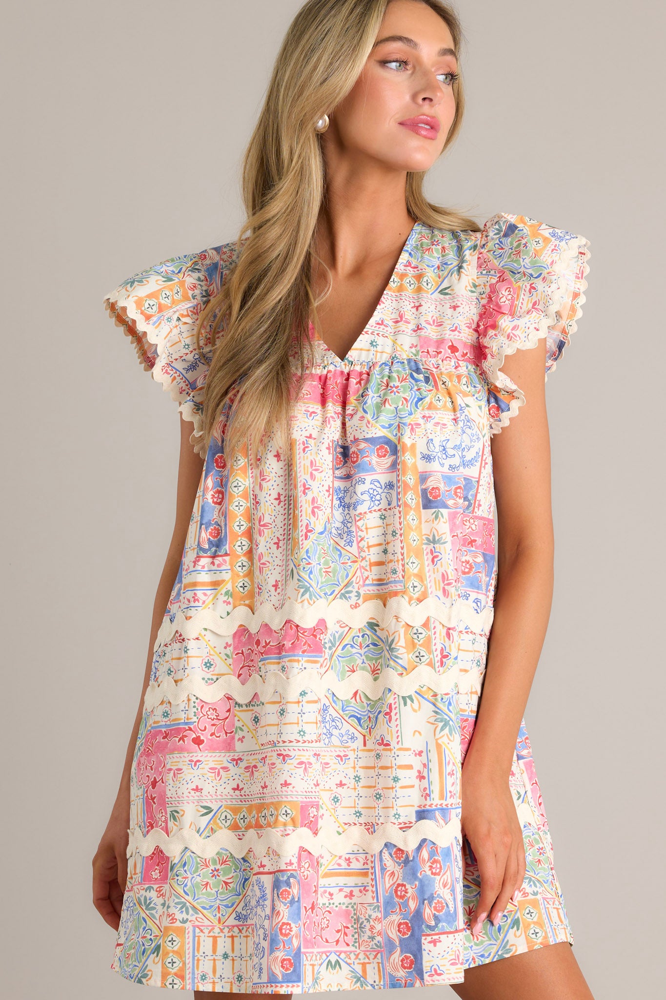 Front angled view of a patchwork mini dress featuring a v-neckline, a unique patchwork design, functional hip pockets, ricrac detailing, and ricrac trim flutter sleeves