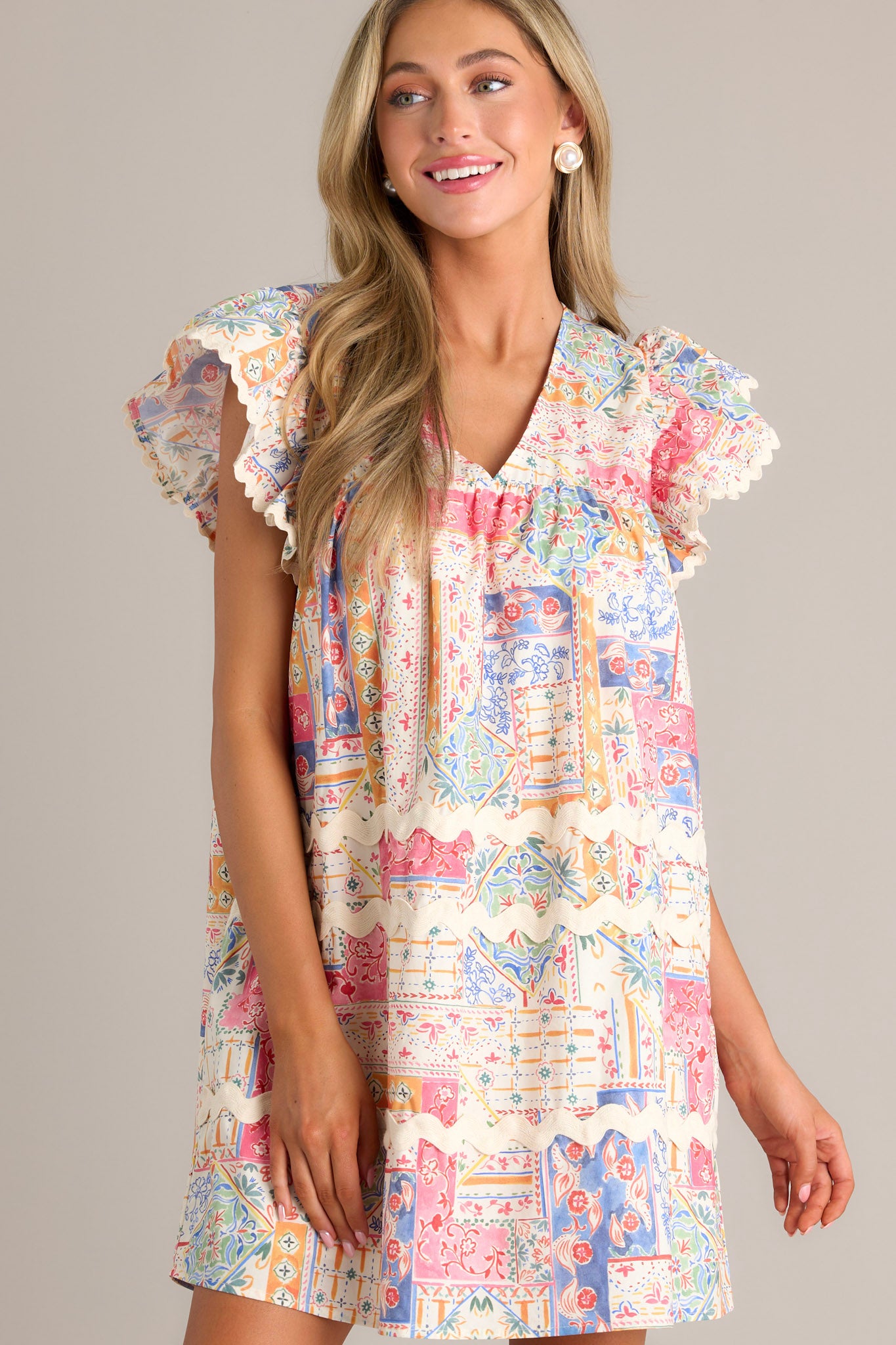 Front view of a patchwork mini dress featuring a v-neckline, a unique patchwork design, functional hip pockets, ricrac detailing, and ricrac trim flutter sleeves.