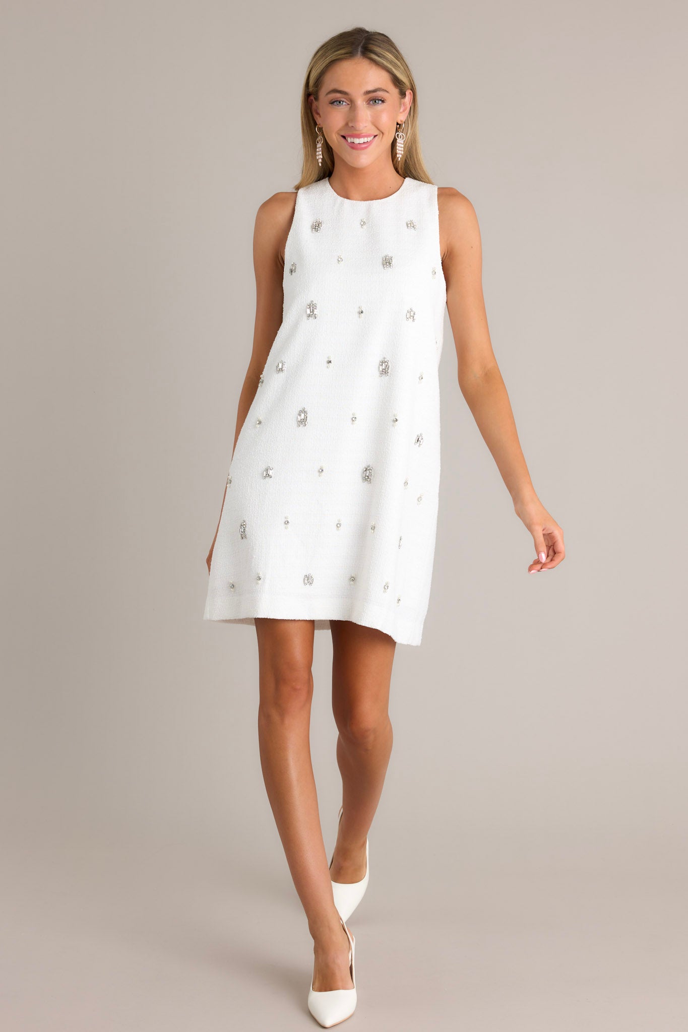 This white mini dress features a crew neckline, a straight silhouette, evenly spaced rhinestone embellishments, and a discrete zipper down the back.