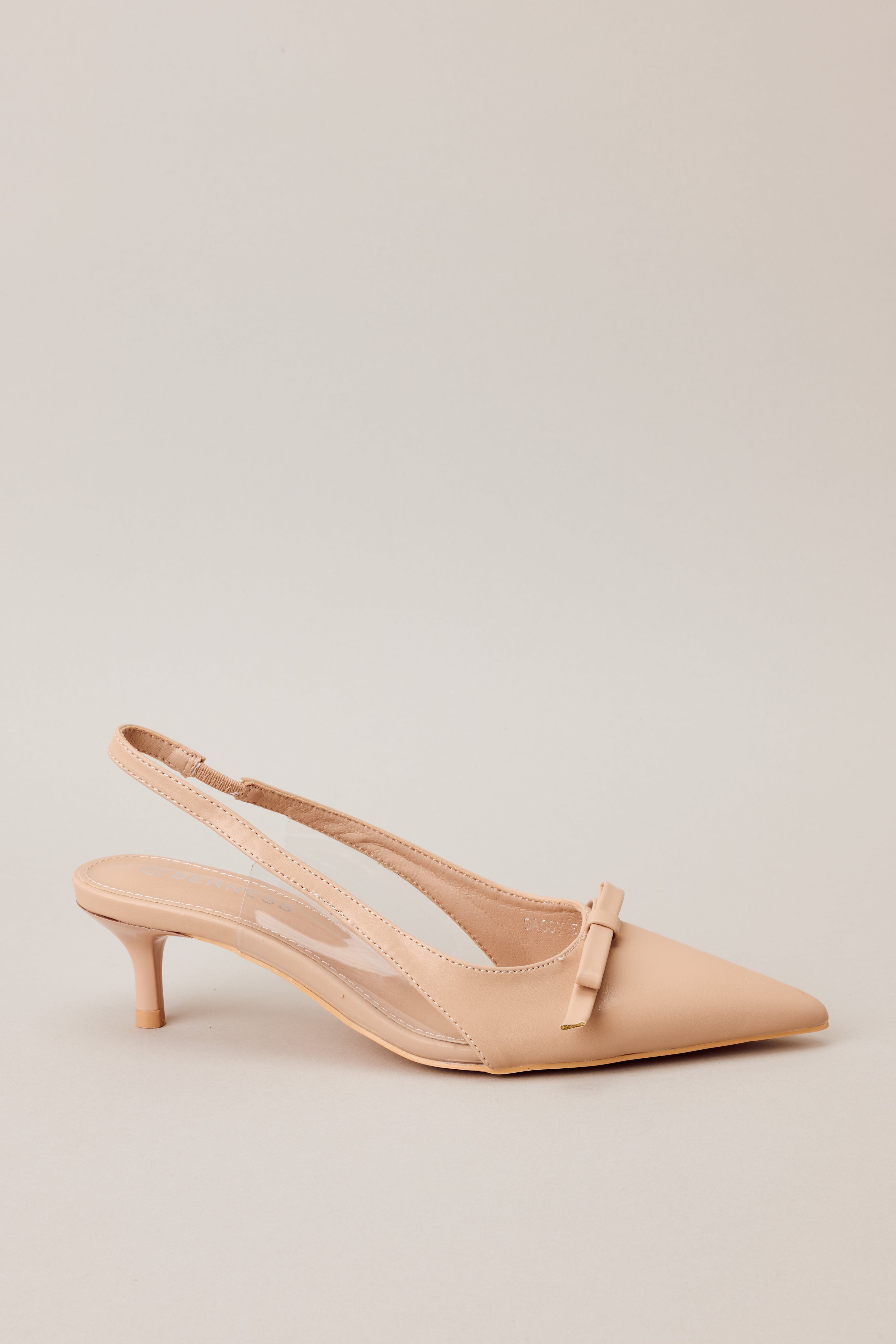 Side view of beige kitten heels showcasing the pointed closed toe, delicate bow detailing, strap around the back of the foot, clear siding for extra support, and extremely short heel.