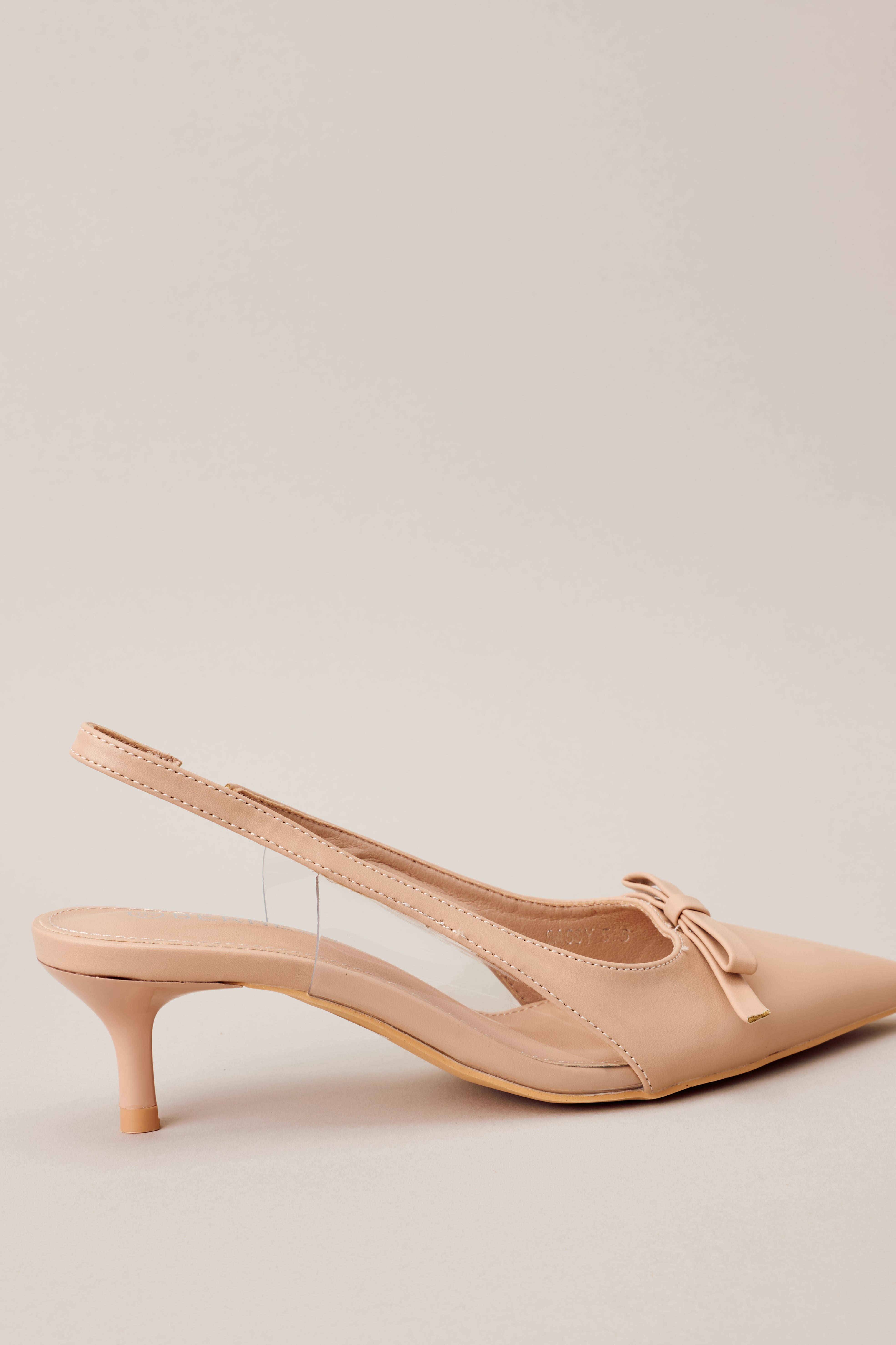 Close-up Side view of beige kitten heels showcasing the pointed closed toe, delicate bow detailing, strap around the back of the foot, clear siding for extra support, and extremely short heel.