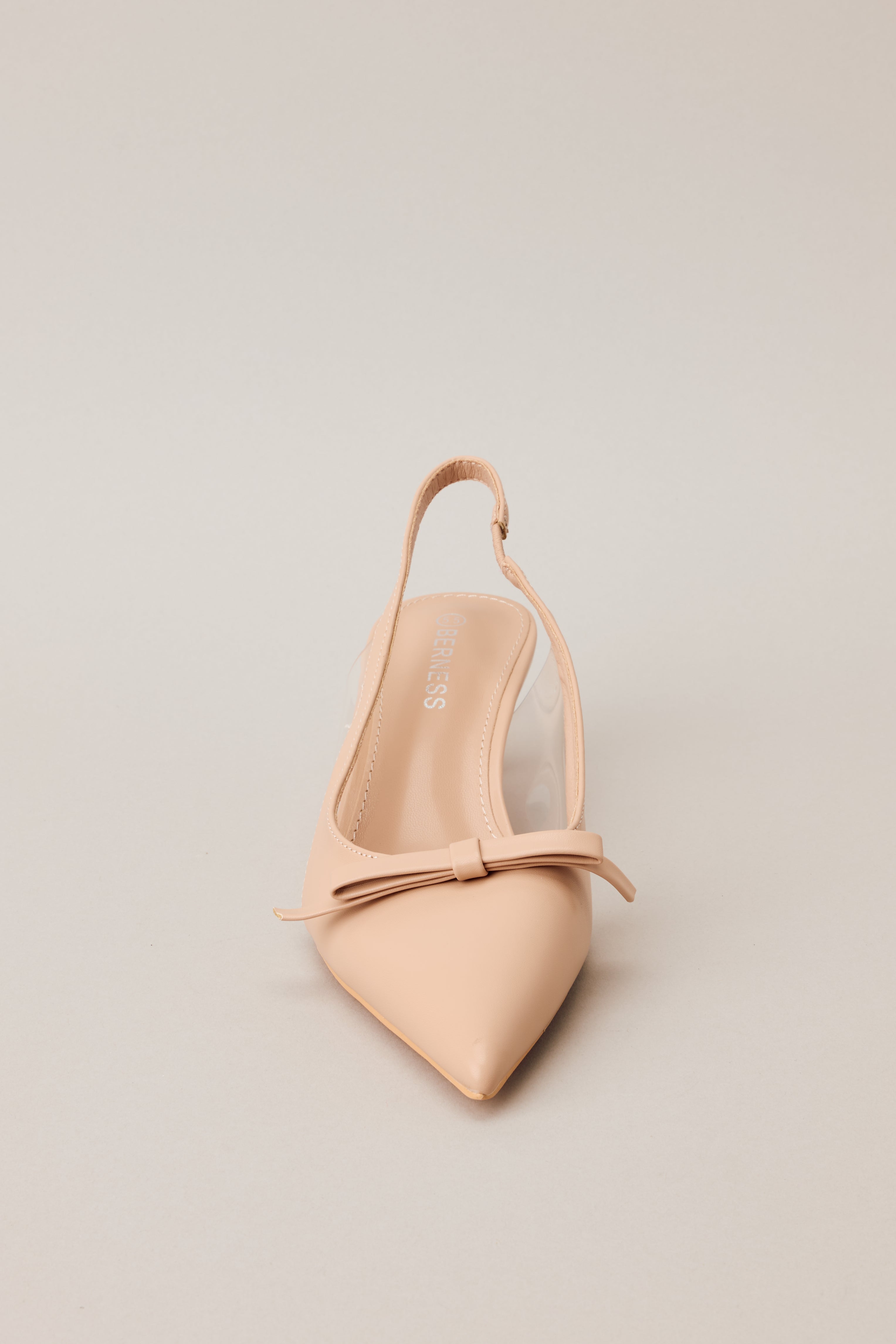 Front view of beige kitten heels featuring a pointed closed toe, delicate bow detailing, a strap around the back of the foot, clear siding for extra support, and an extremely short heel.