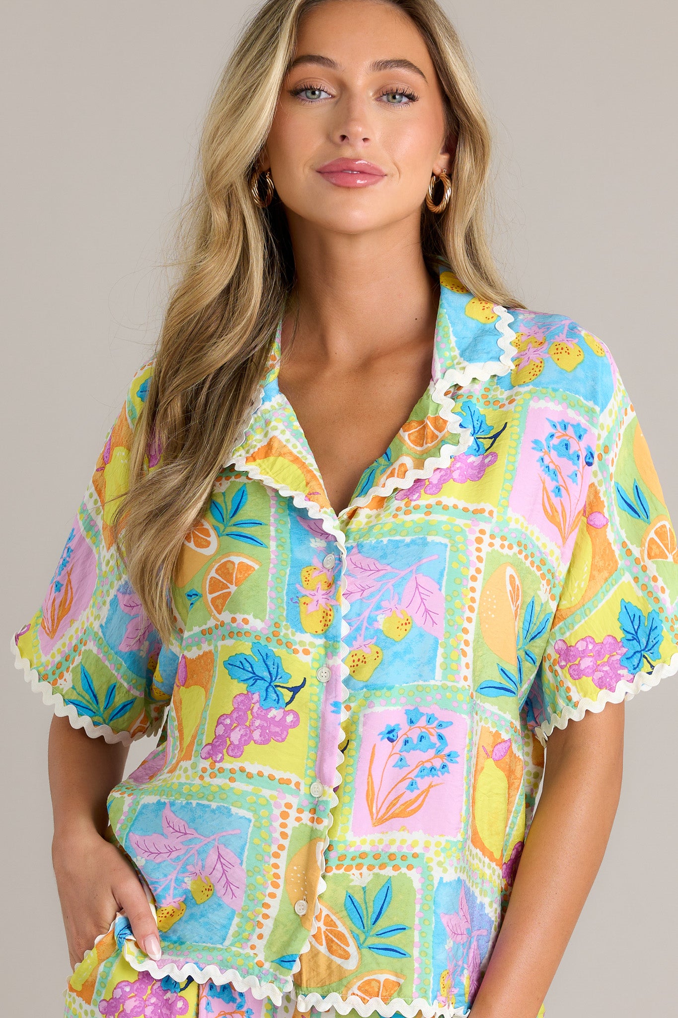 Front angled view of a tropical ricrac top featuring a notched lapel collared neckline, a functional button front, a unique print, and contrasting ricrac trim