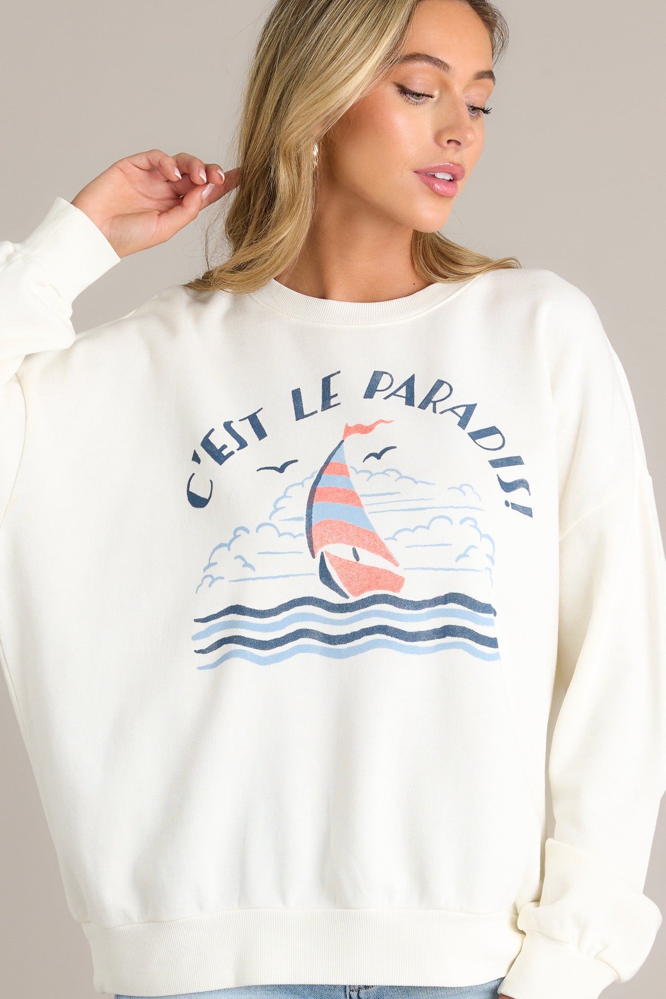 Side view of a white sweatshirt showcasing the crew neckline, sailboat themed graphic, ribbed cuffed long sleeves, and ribbed hemline.