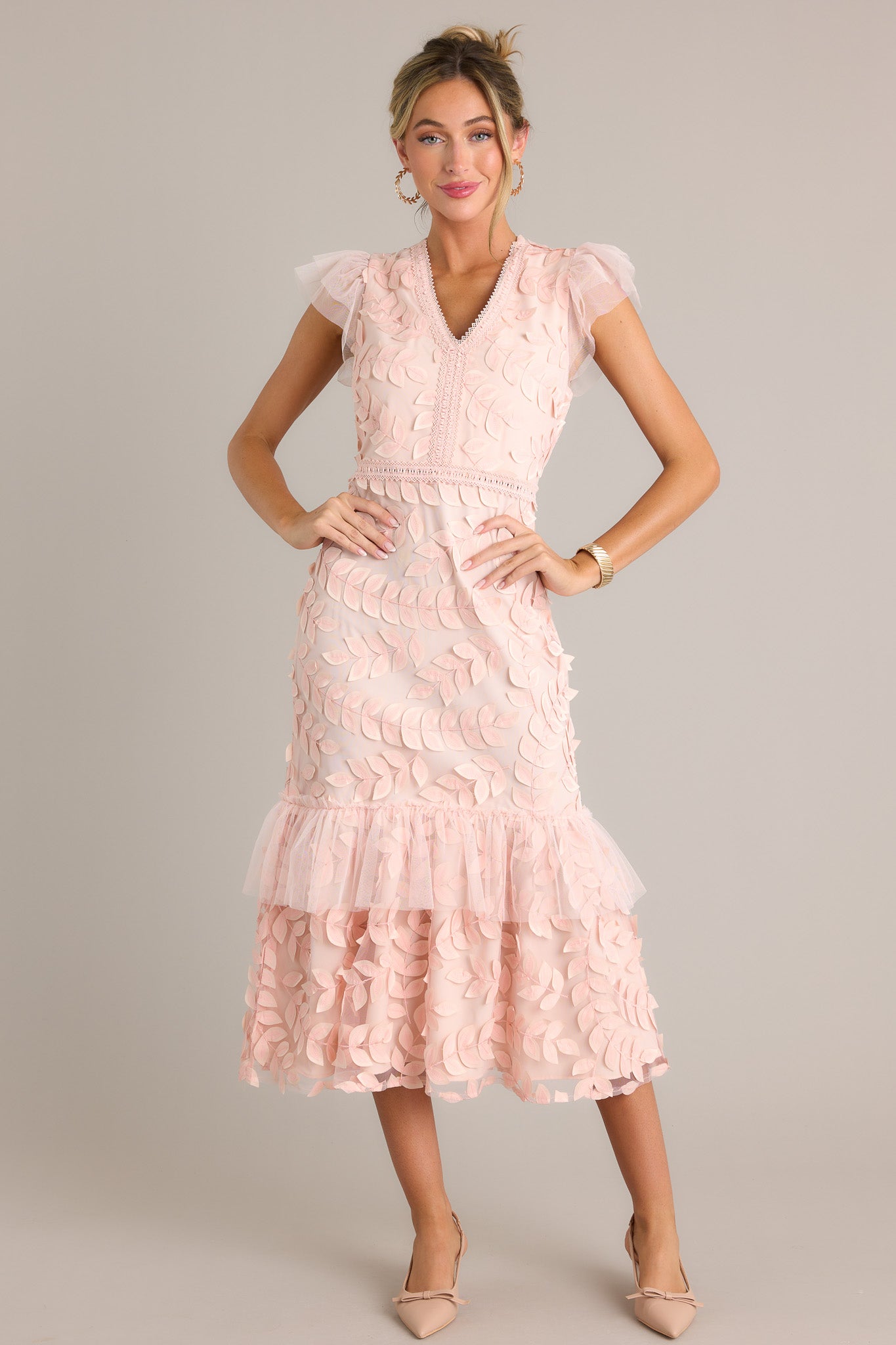 Full length view of a light pink midi dress with a v-neckline, a discrete back zipper, embroidered detailing, 3-D petals throughout, a lace tier, and lace short sleeves