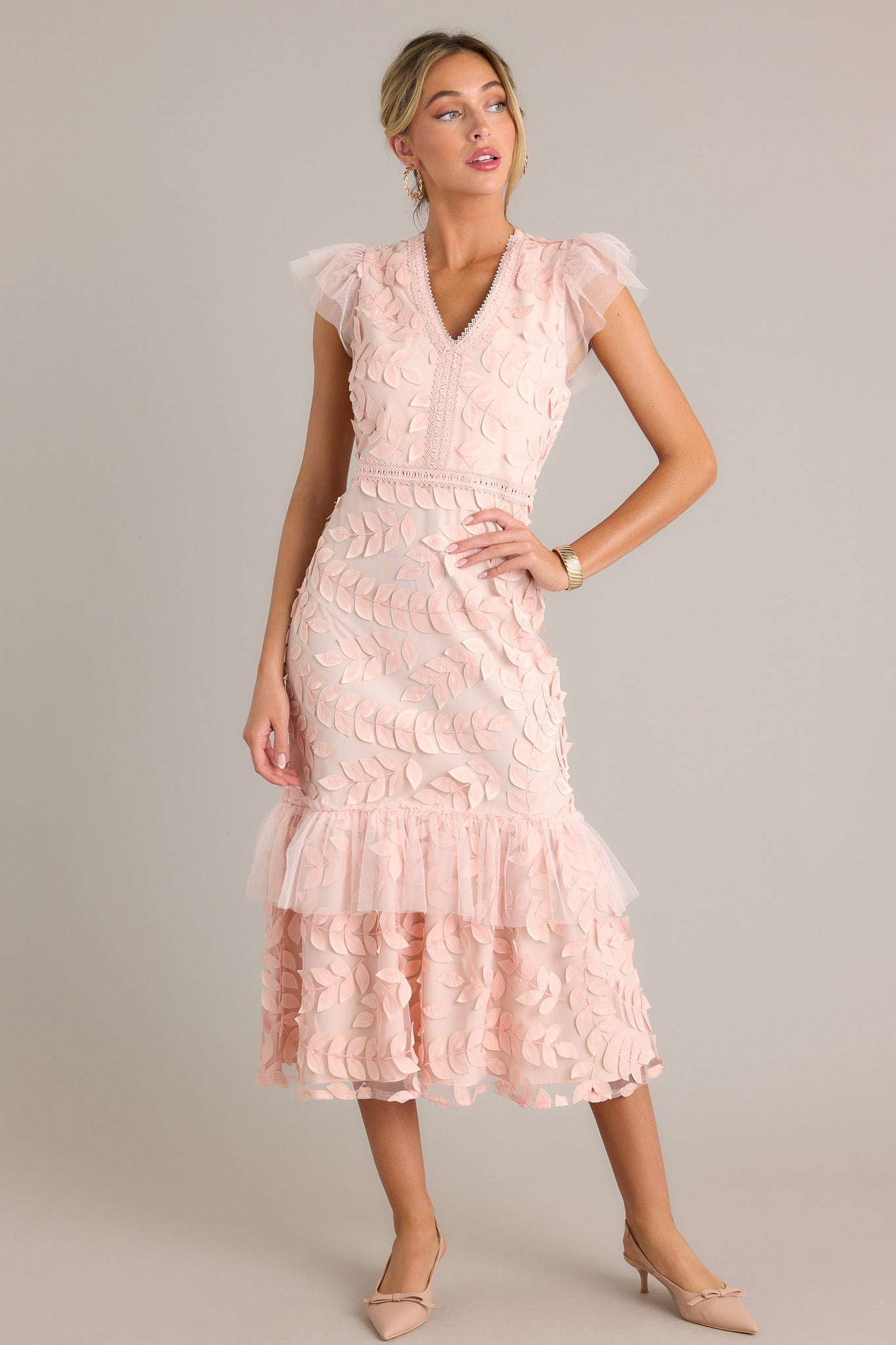 Front angled view of a light pink midi dress featuring a v-neckline, a discrete back zipper, embroidered detailing, 3-D petals throughout, a lace tier, and lace short sleeves