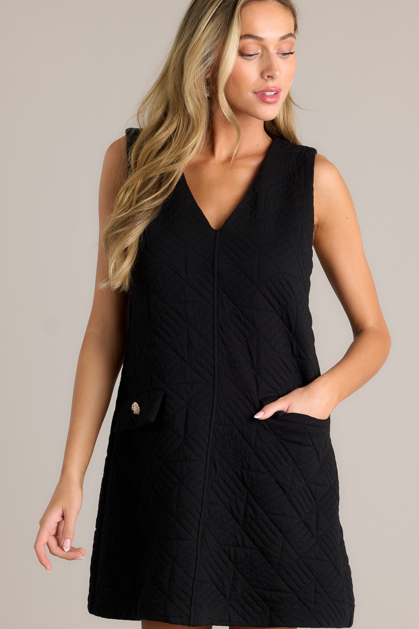 Front angled view of a black mini dress featuring a v-neckline, a quilted fabric, functional pockets with faux buttons, and a sleeveless design