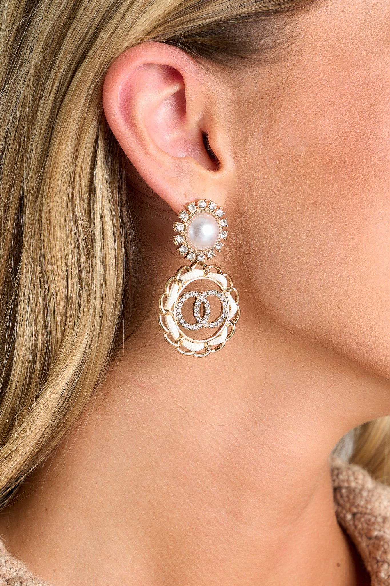 Close-up view of these gold earrings that feature a pearl and rhinestone stud, a circle chain pendant with an ivory leather strap woven through it, and a double circle centerpiece with rhinestone accents