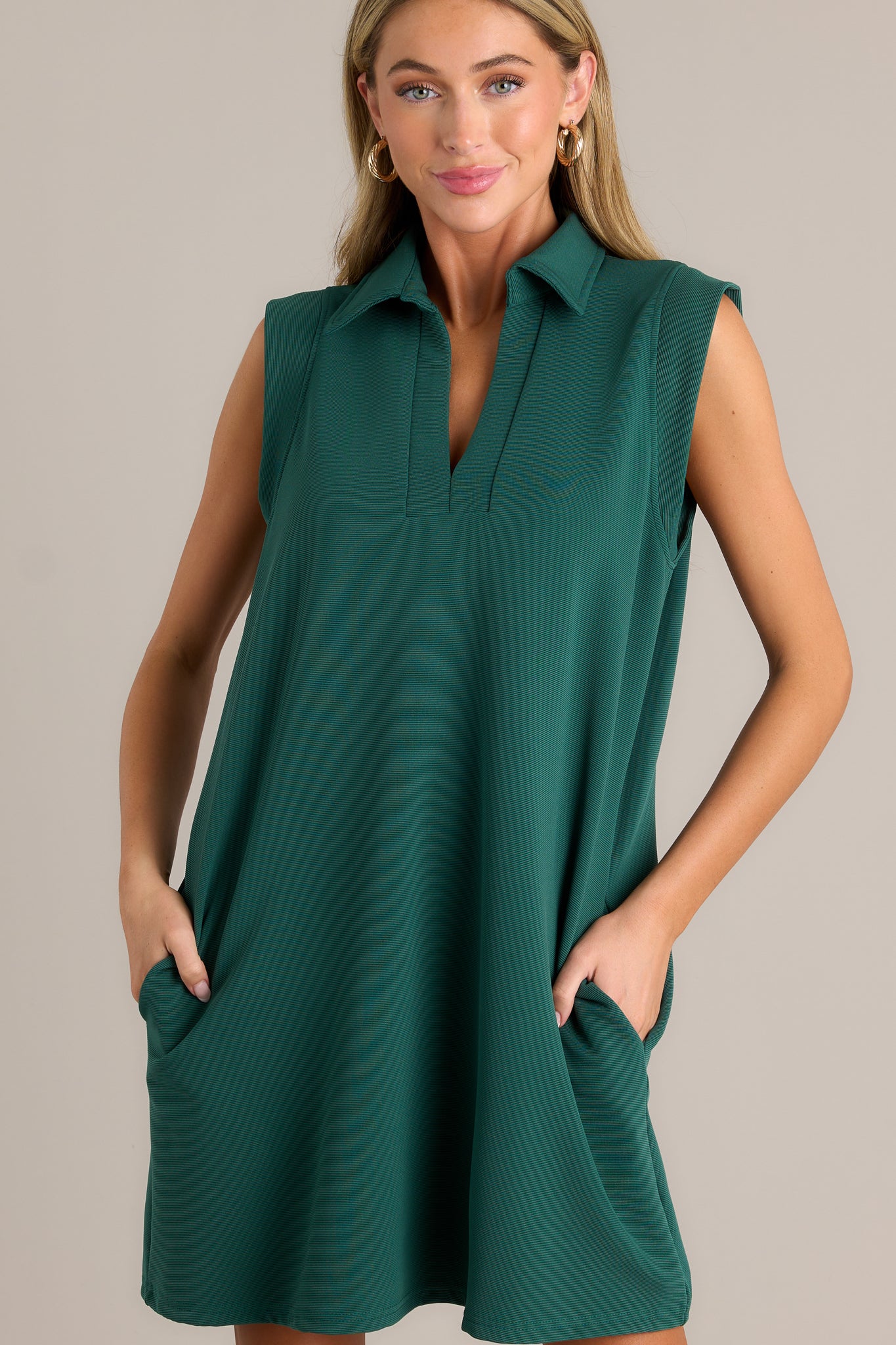 Front view of a green mini dress featuring a collared v-neckline, functional hip pockets, a relaxed fit, and a sleeveless design.