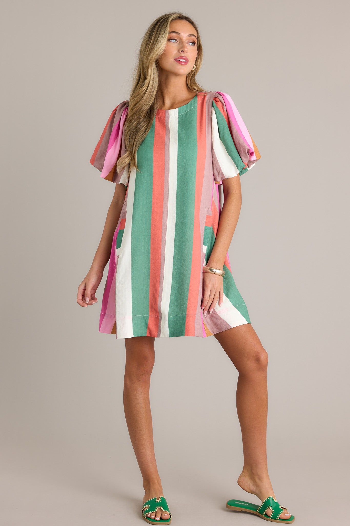 Front angled view of a stripe mini dress featuring a rounded neckline, vertical multi-colored stripes, functional hip pockets, a thick hemline, and elastic cuffed puff sleeves