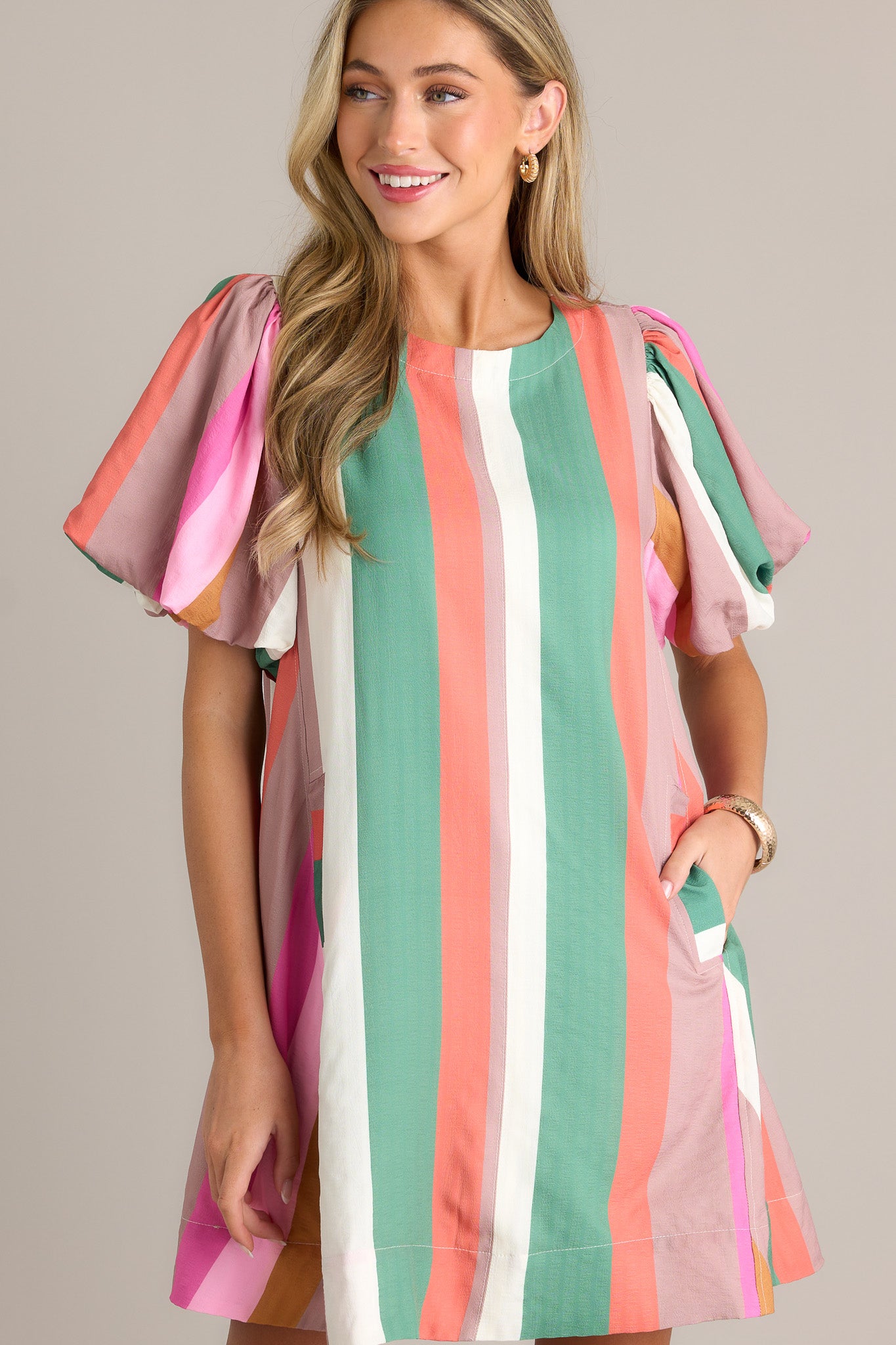 Front view of a stripe mini dress featuring a rounded neckline, vertical multi-colored stripes, functional hip pockets, a thick hemline, and elastic cuffed puff sleeves.