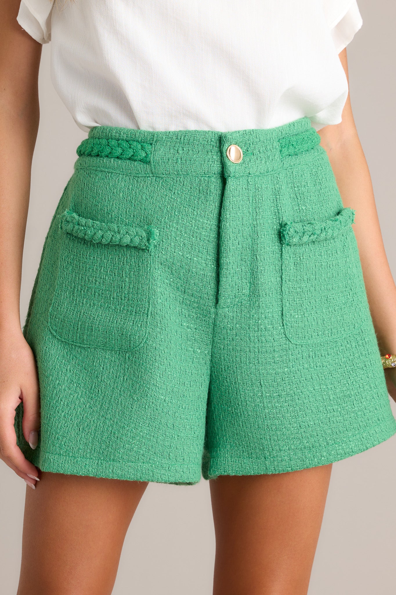 Front view of kelly green tweed shorts featuring a high waisted design, braided waist detailing, a gold accent button, functional front pockets, and a discrete zipper.