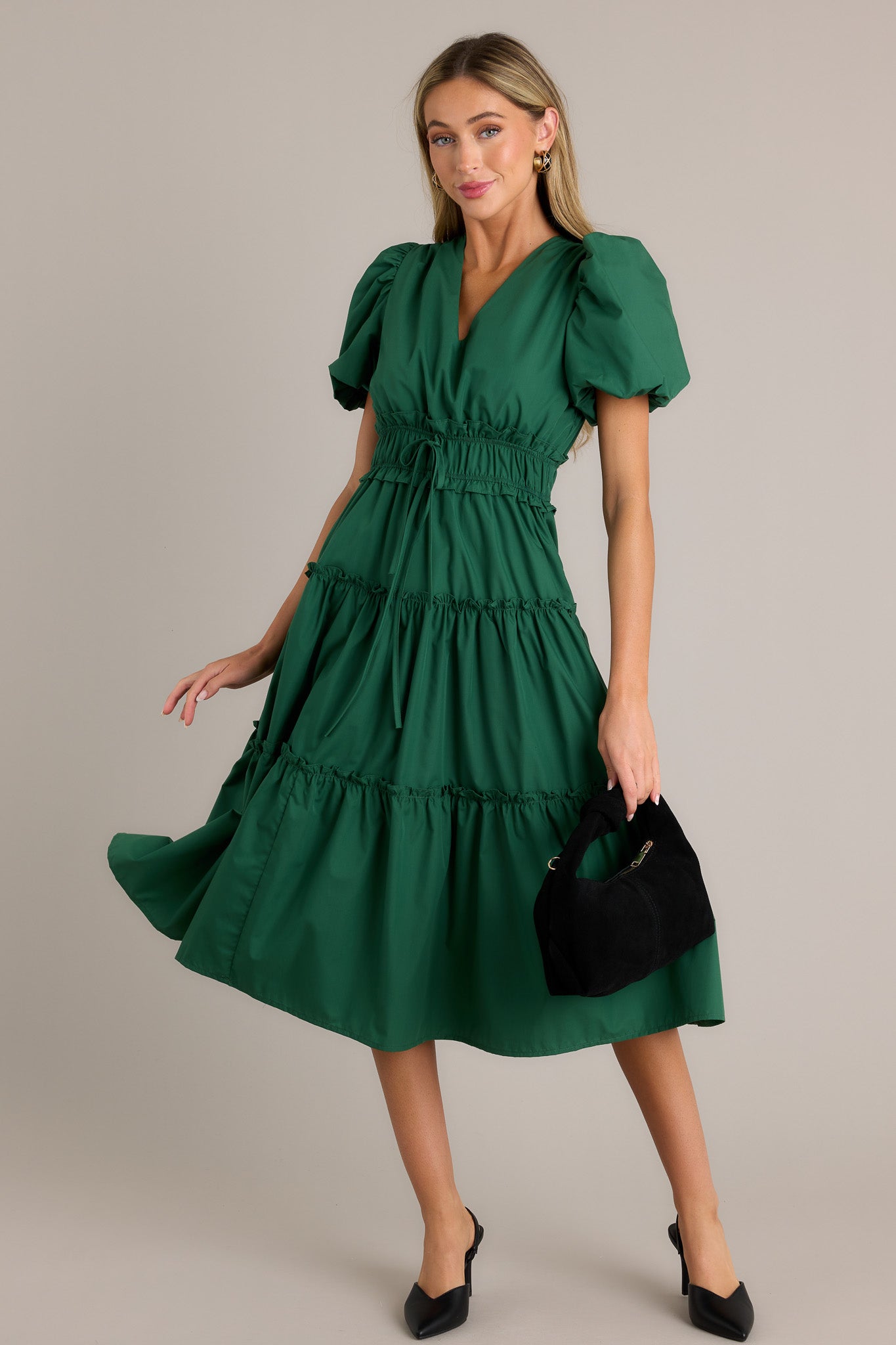 This green midi dress features a v-neckline, a ruffled elastic waistband, self-tie waist features, functional hip pockets, a tiered design, and short puffed sleeves.