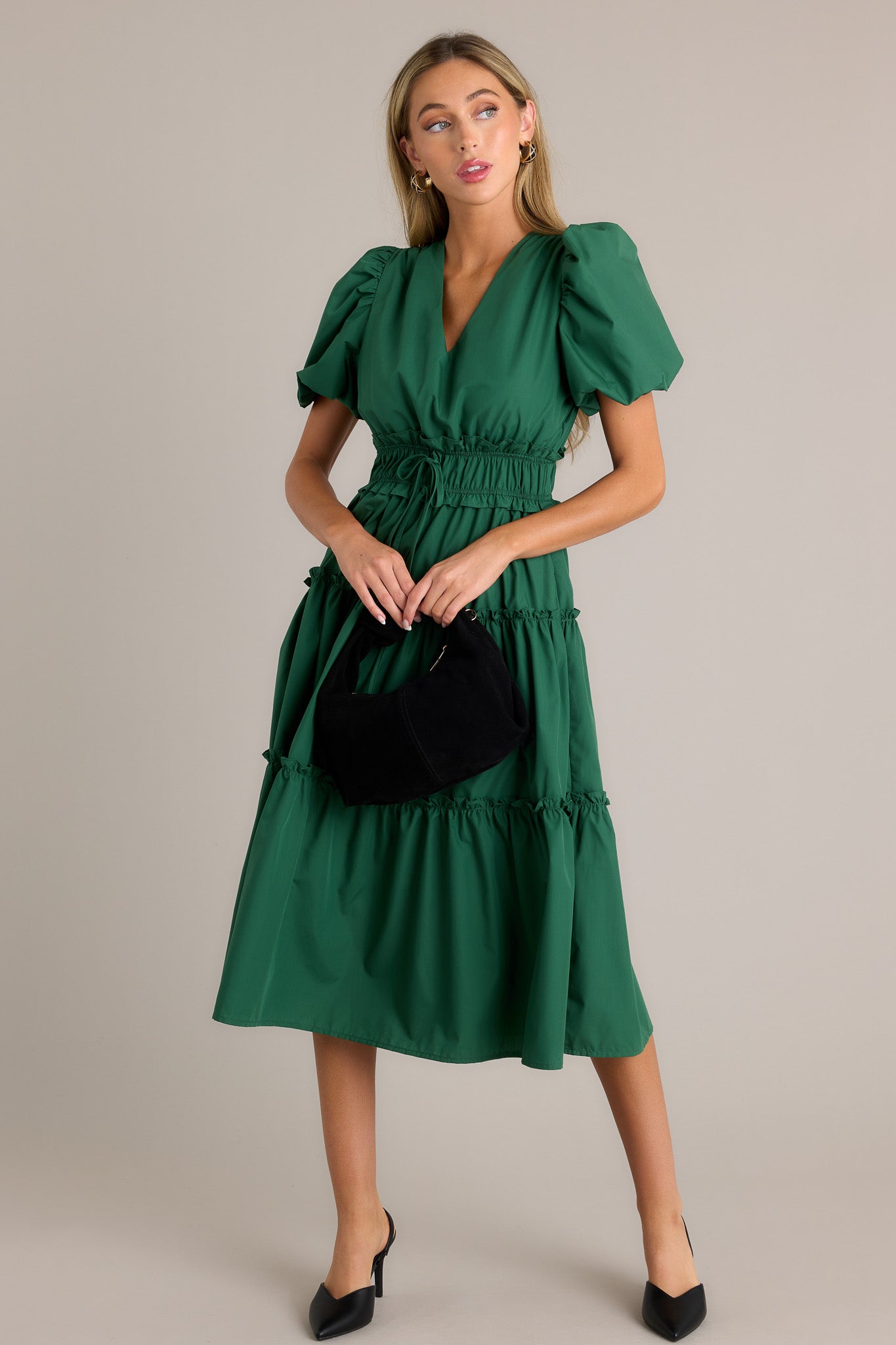 Front angled view of a green midi dress featuring a v-neckline, a ruffled elastic waistband, self-tie waist features, functional hip pockets, a tiered design, and short puffed sleeves